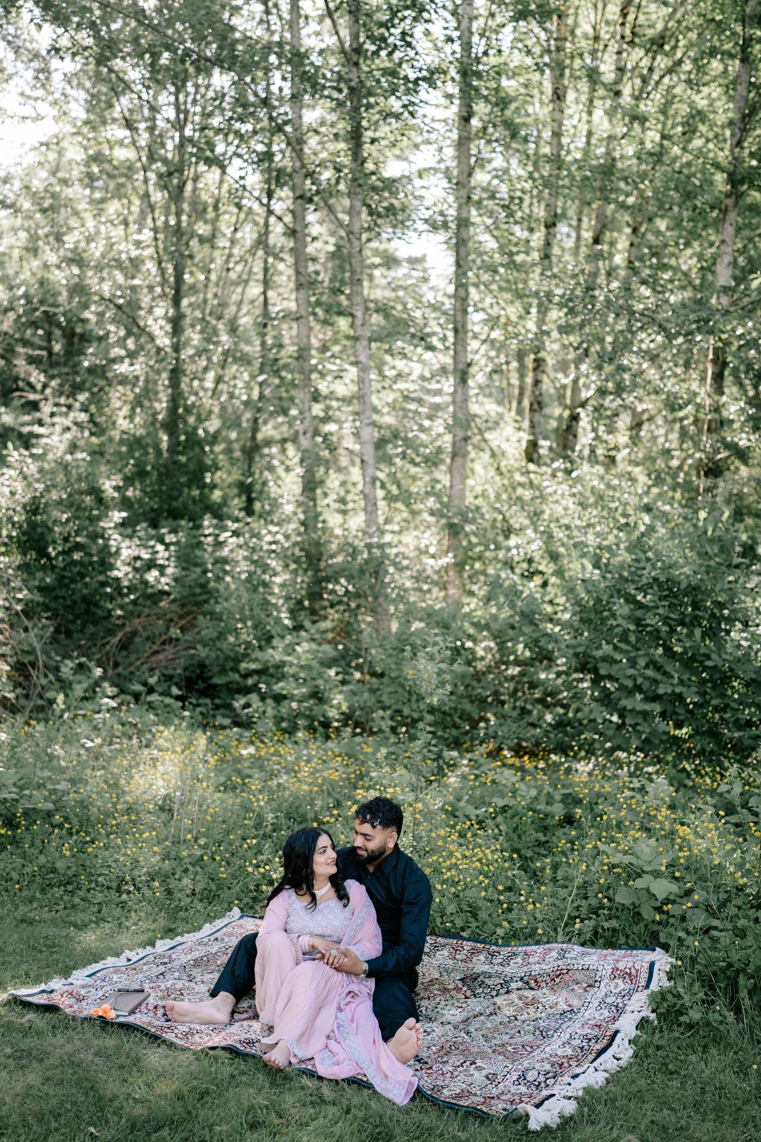 Couple relaxes on patterned blanket during outdoor photoshoot in wooded setting.