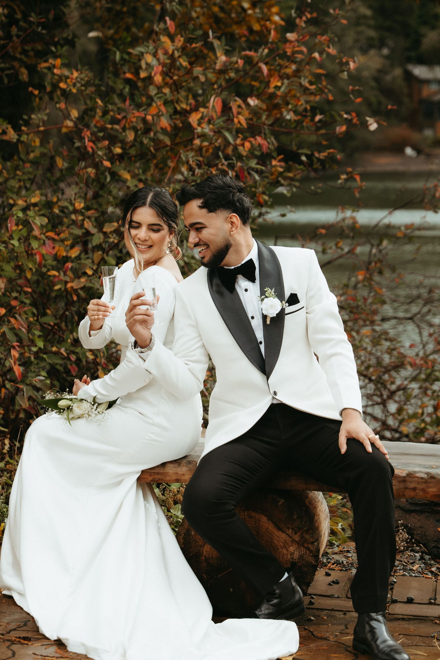 Elegant couple in white wedding attire share drinks while sitting outdoors among autumn foliage.