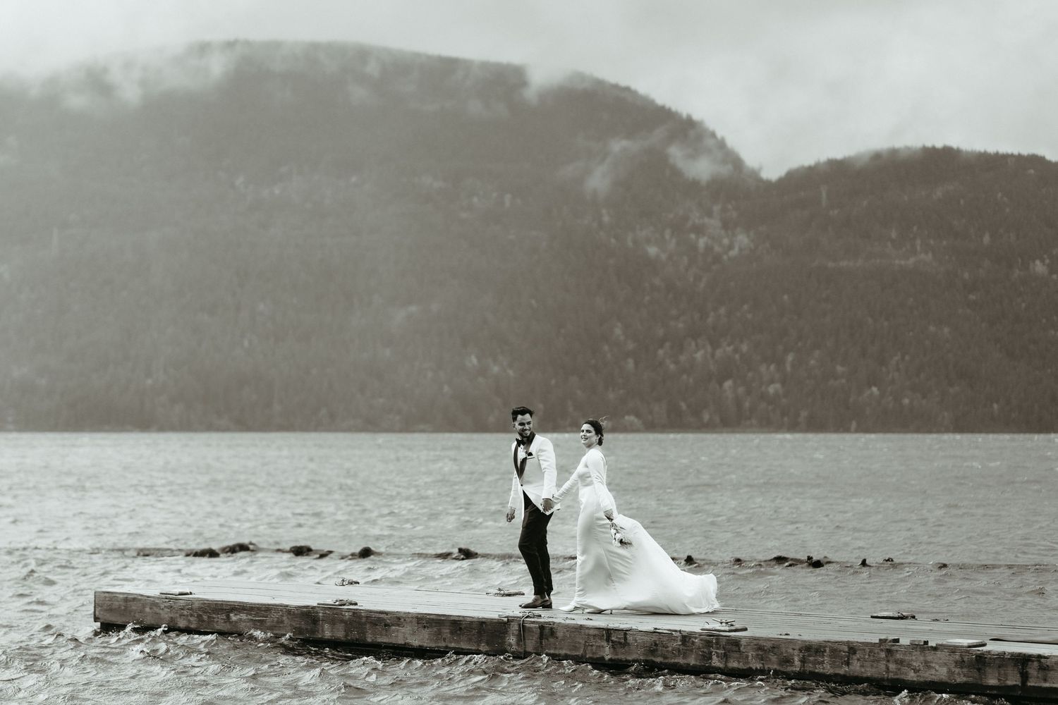 A couple in wedding attire stands on a stone pier overlooking a misty lake and mountains in black and white.