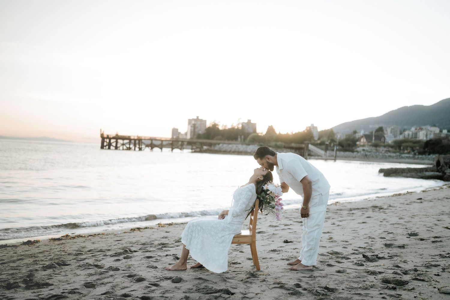 A couple in white outfits dances together on a scenic beach at sunset with a pier in the background.