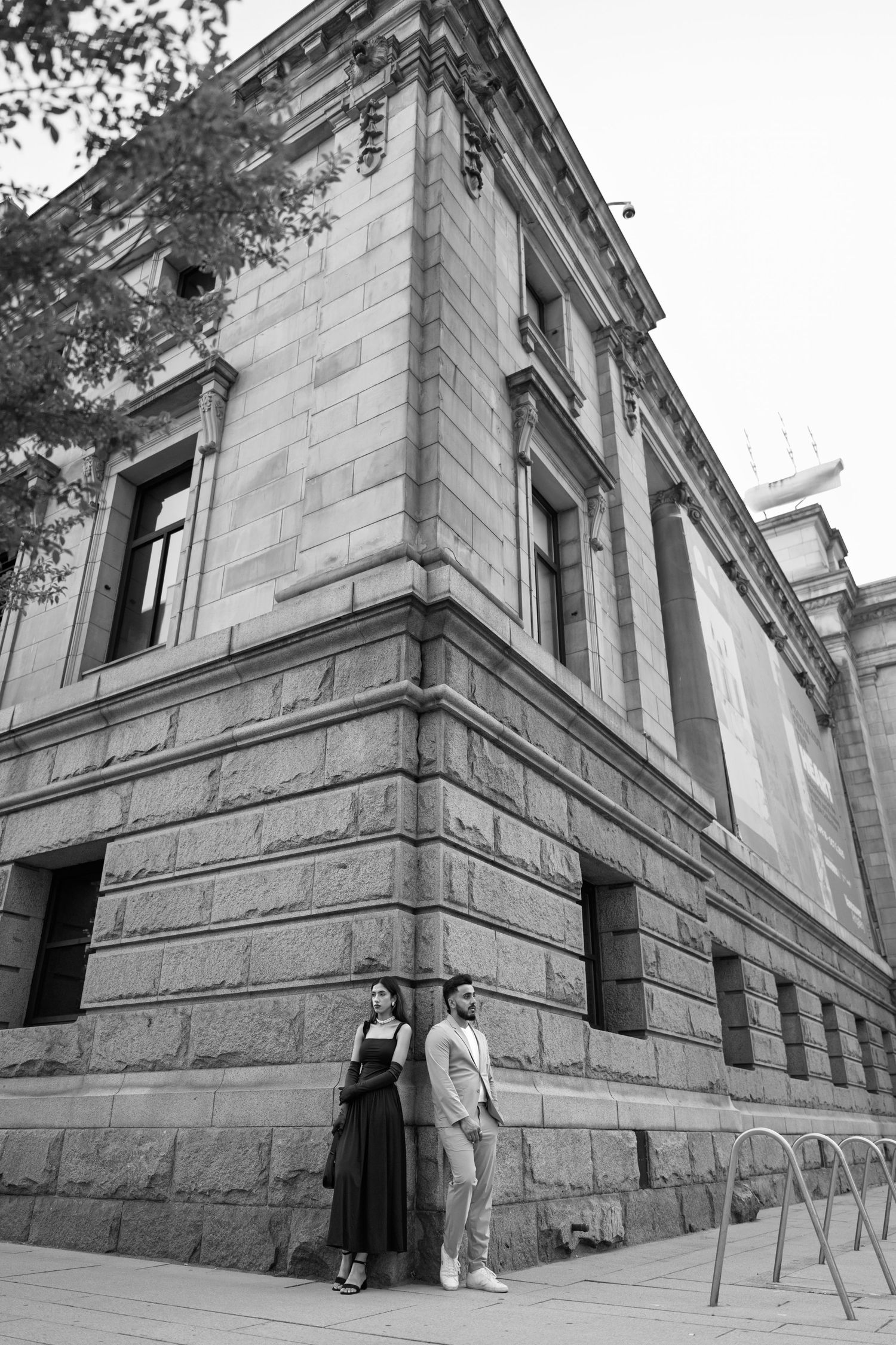 Black and white architectural photo showing two people standing at the corner of a grand stone building with classical details.