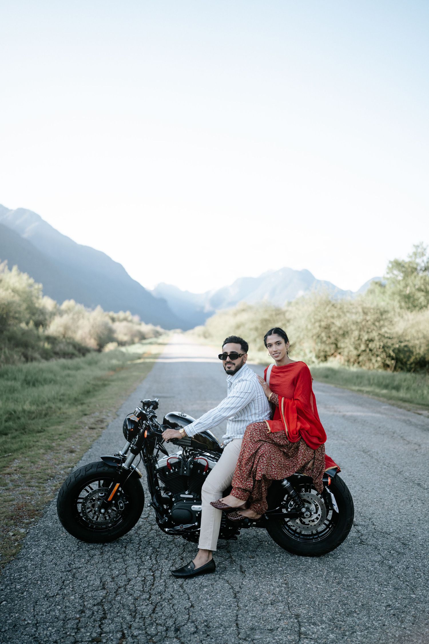 Couple poses with vintage motorcycle on mountain road against scenic backdrop of rugged peaks and greenery.