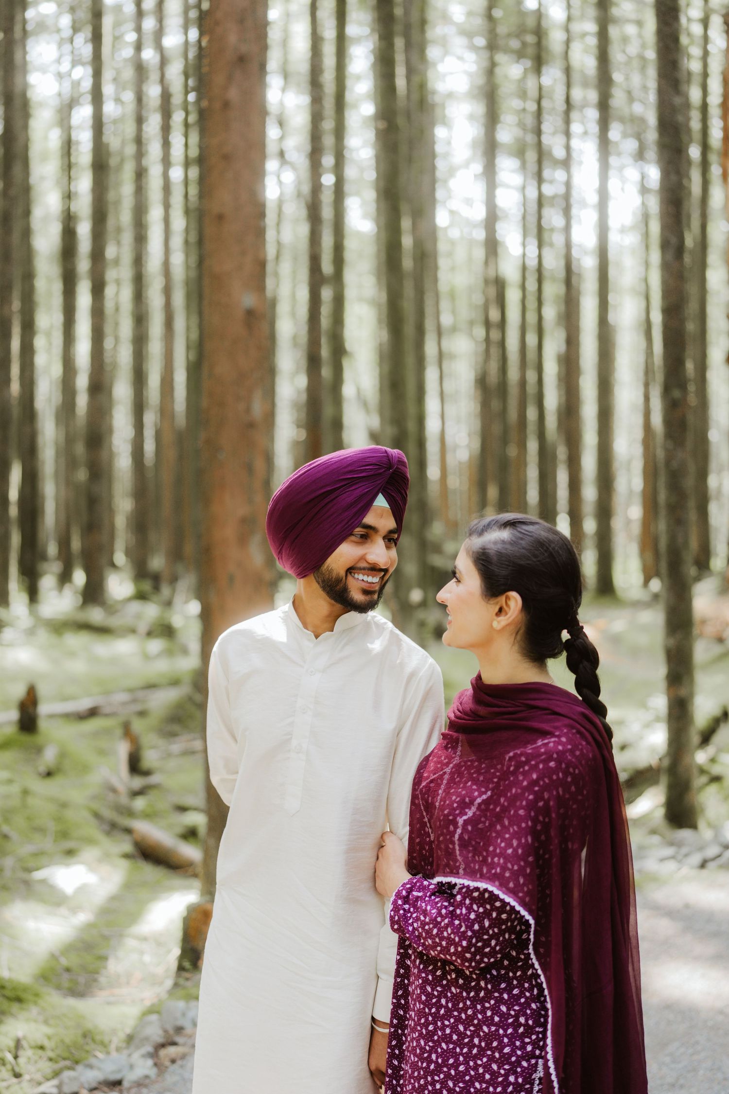 Smiling Sikh man in purple turban and white kurta gazing at woman in purple salwar suit in forest.