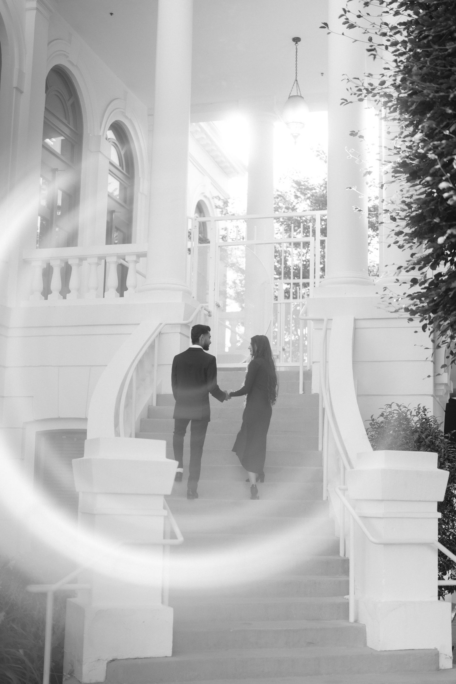 Black and white image of a couple holding hands on grand white outdoor staircase with columns.