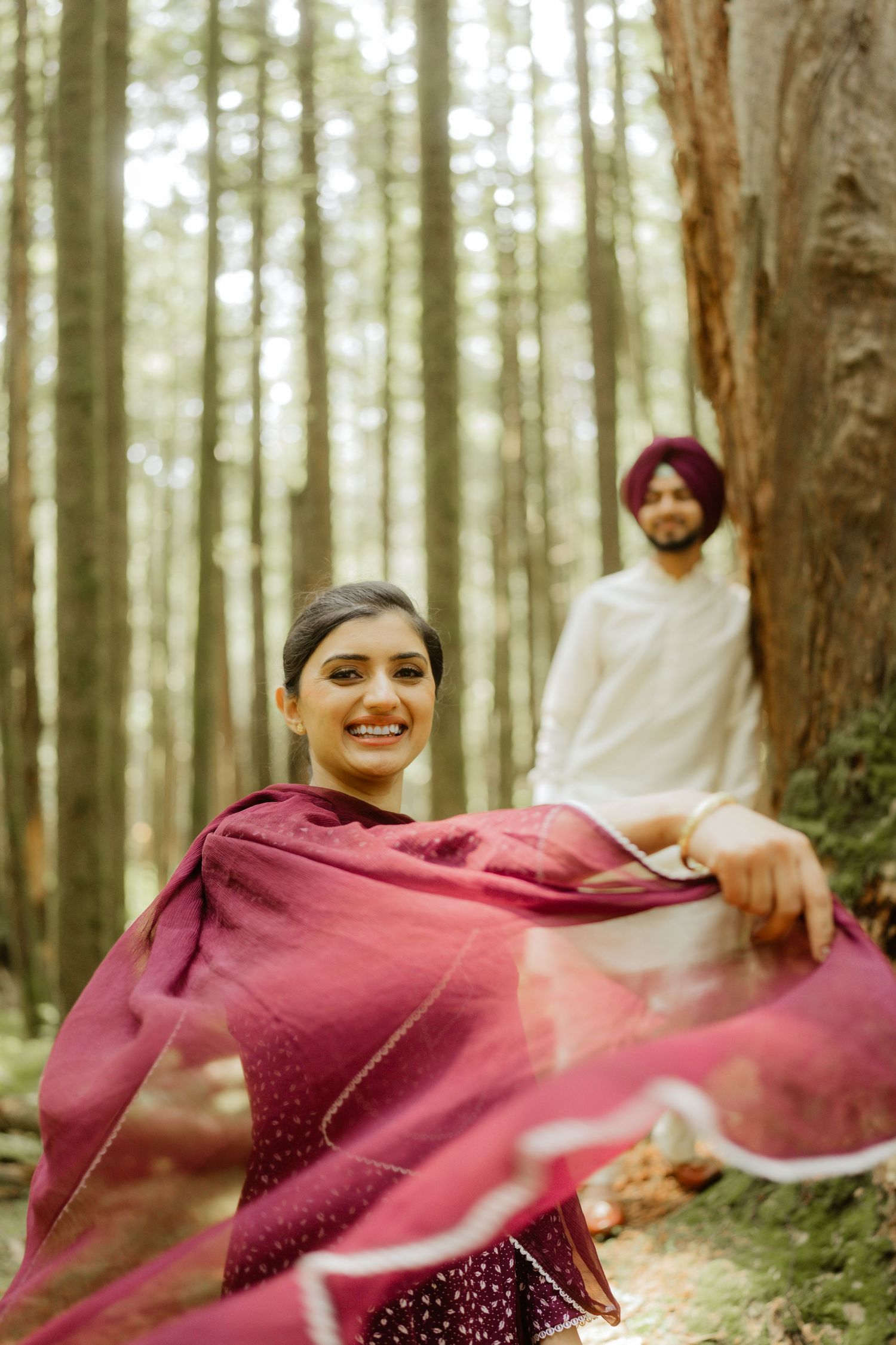 A smiling woman in a flowing burgundy dupatta poses in a forest, with a Sikh man in white sherwani in the background.