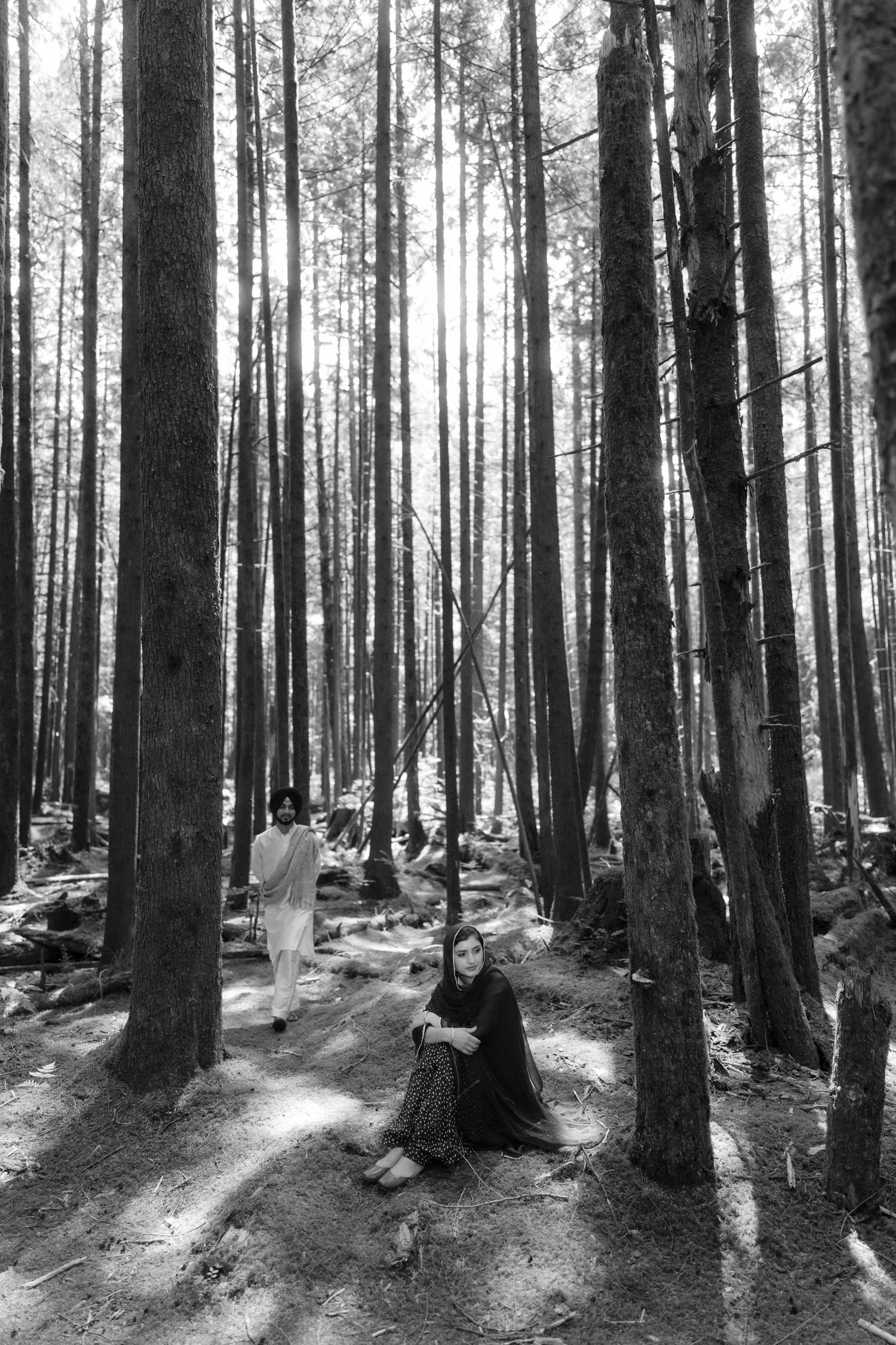 Black and white photo of a Sikh couple in a tall pine forest, woman seated on ground, man walking behind.