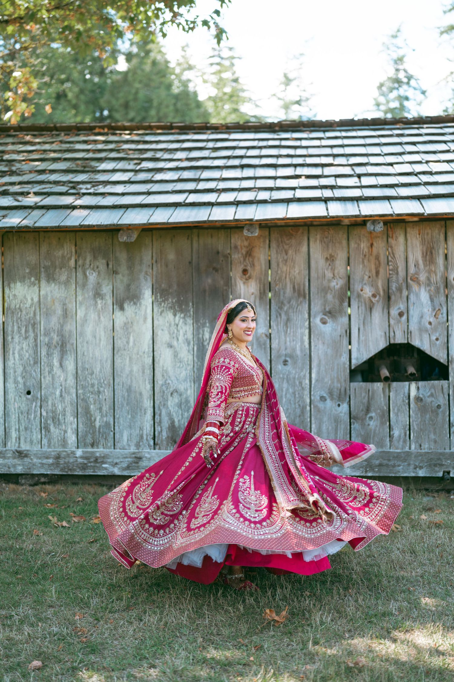 Bride twirling in vibrant pink embroidered lehenga outdoors near rustic wooden barn.