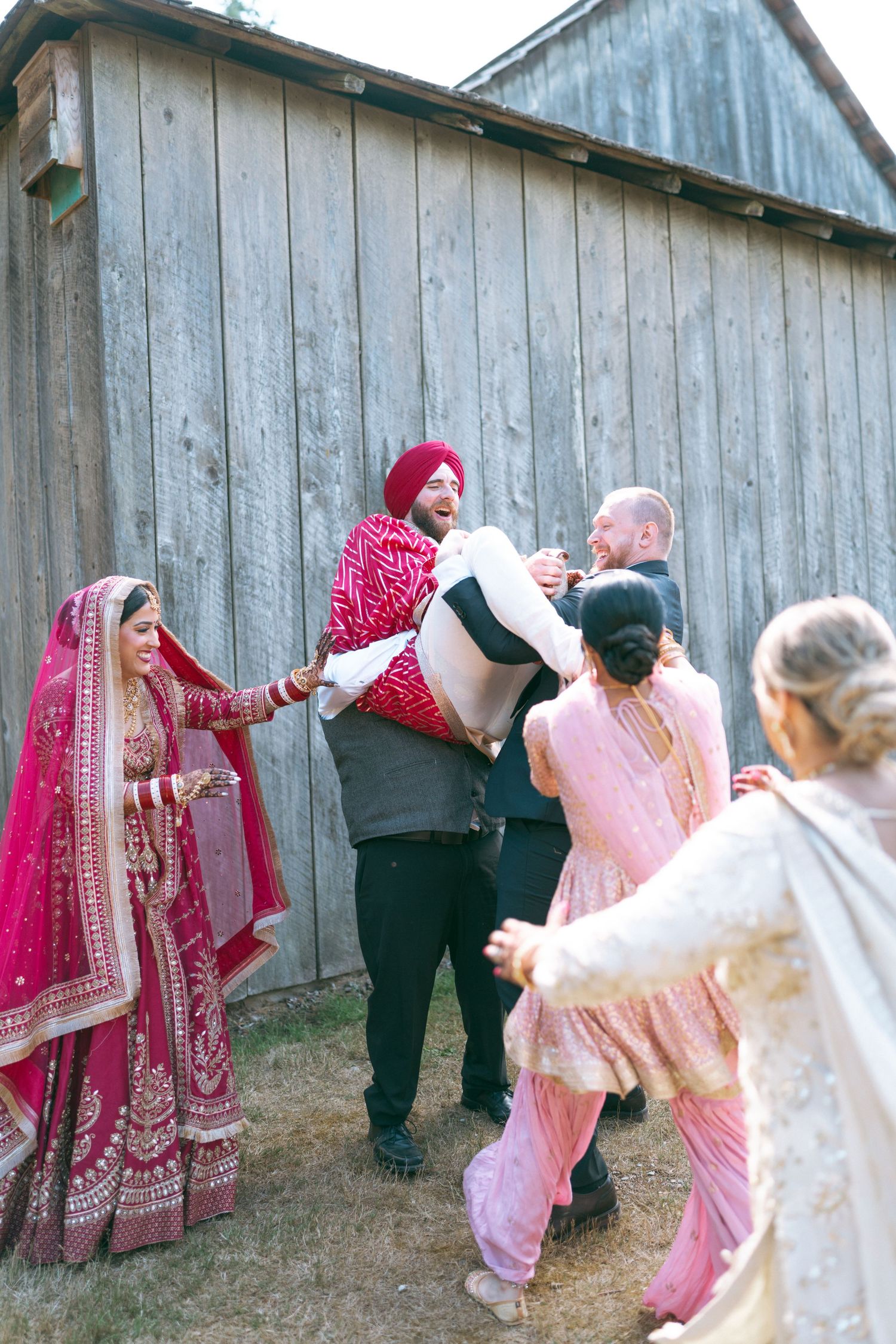 Sikh groom in red turban lifted by groomsmen while bride in red lehenga and guests celebrate at outdoor wedding.