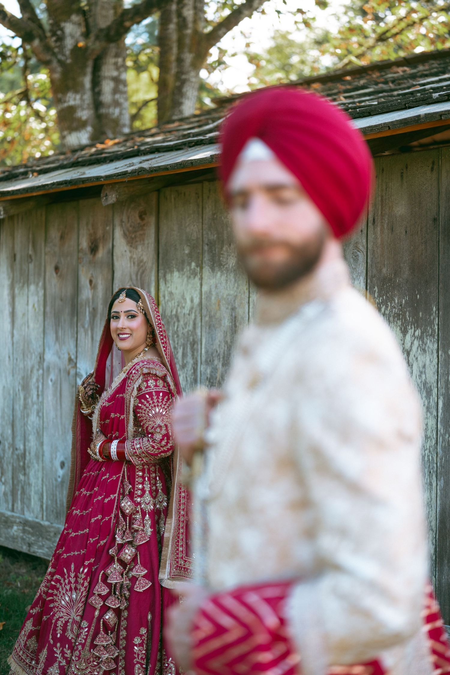 Sikh bride in red lehenga stands behind groom in cream sherwani and red turban during traditional wedding ceremony.