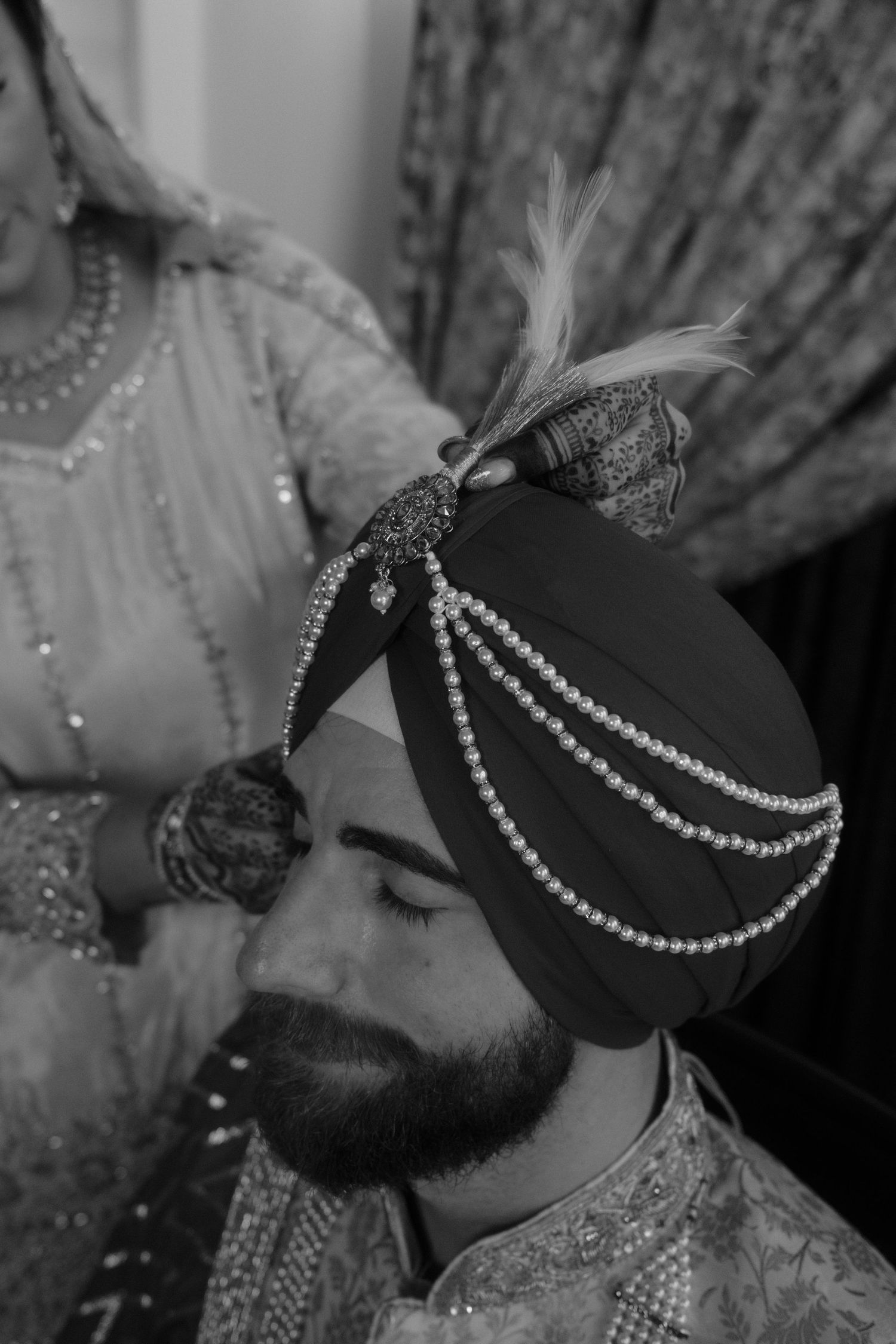 Black and white photo of a Sikh groom having his decorated turban adorned with pearl chains and feather during wedding.