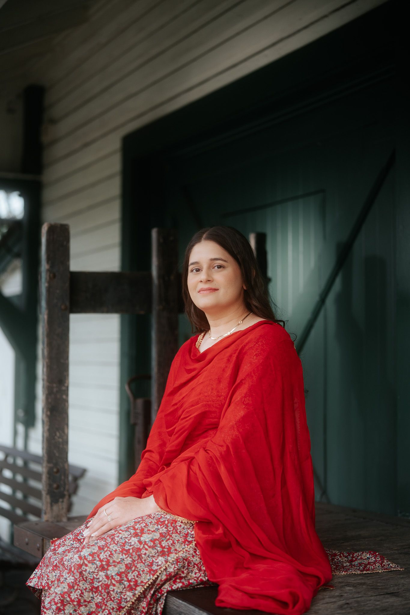 Young woman in red dupatta and floral dress sitting peacefully on a wooden bench outside a heritage building.