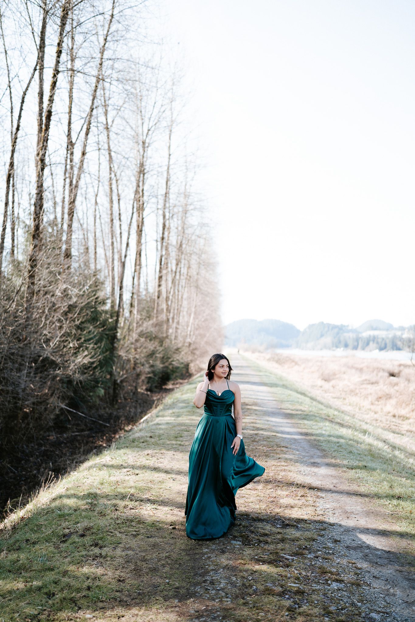 A person in a flowing teal dress poses gracefully along a serene forest path on a misty winter day.