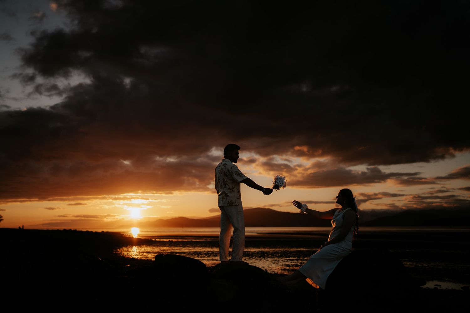 Romantic silhouette of a couple sharing a moment on rocks during a dramatic sunset by the water.