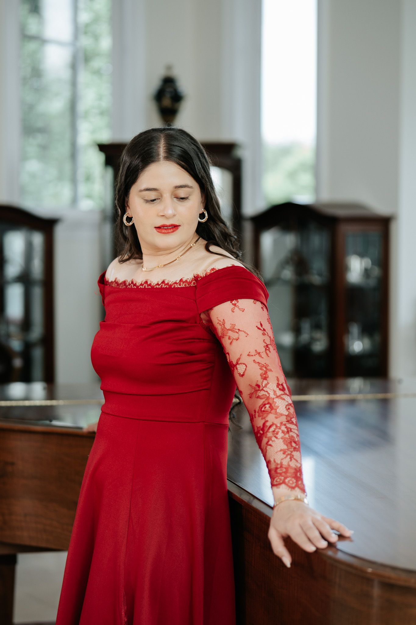 Woman in elegant red off-shoulder gown with lace sleeves posing near a grand piano in a classic interior.