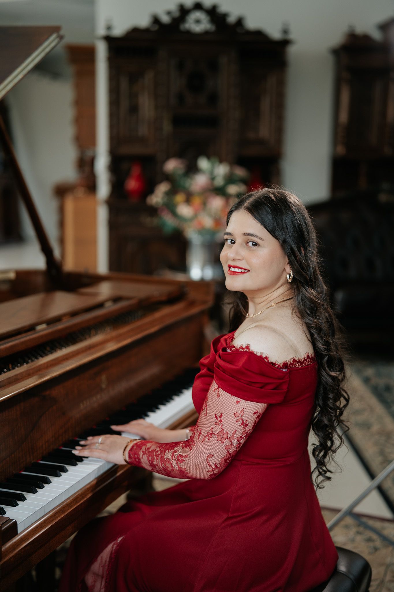 A woman in an elegant red lace off-shoulder gown smiles while playing a grand piano in a sophisticated interior.