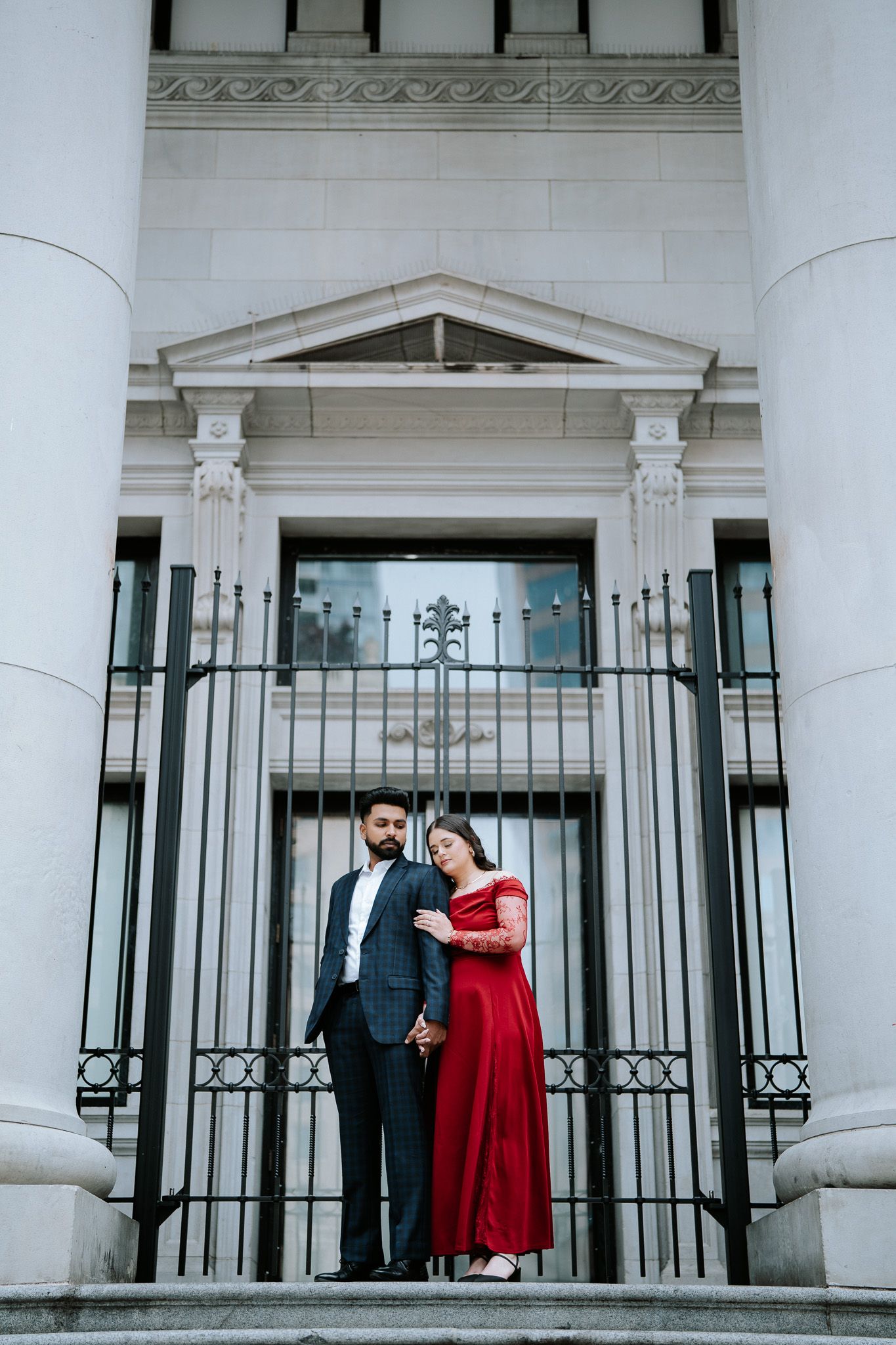 A couple stands on grand white building steps, the woman in a red gown holding hands with a man in a dark suit.
