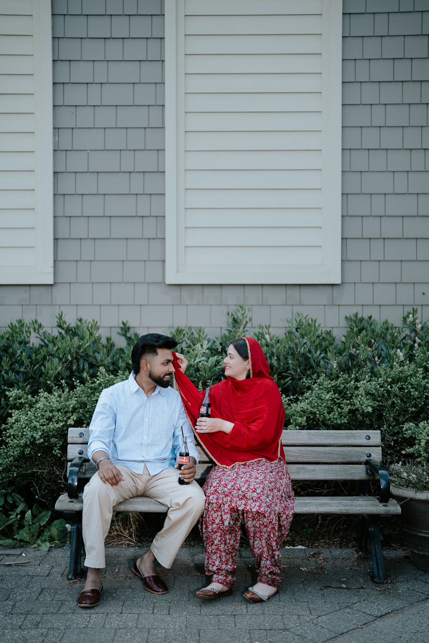 Couple sitting on a bench, woman in red salwar kameez touching man's face, sharing a tender moment outdoors.