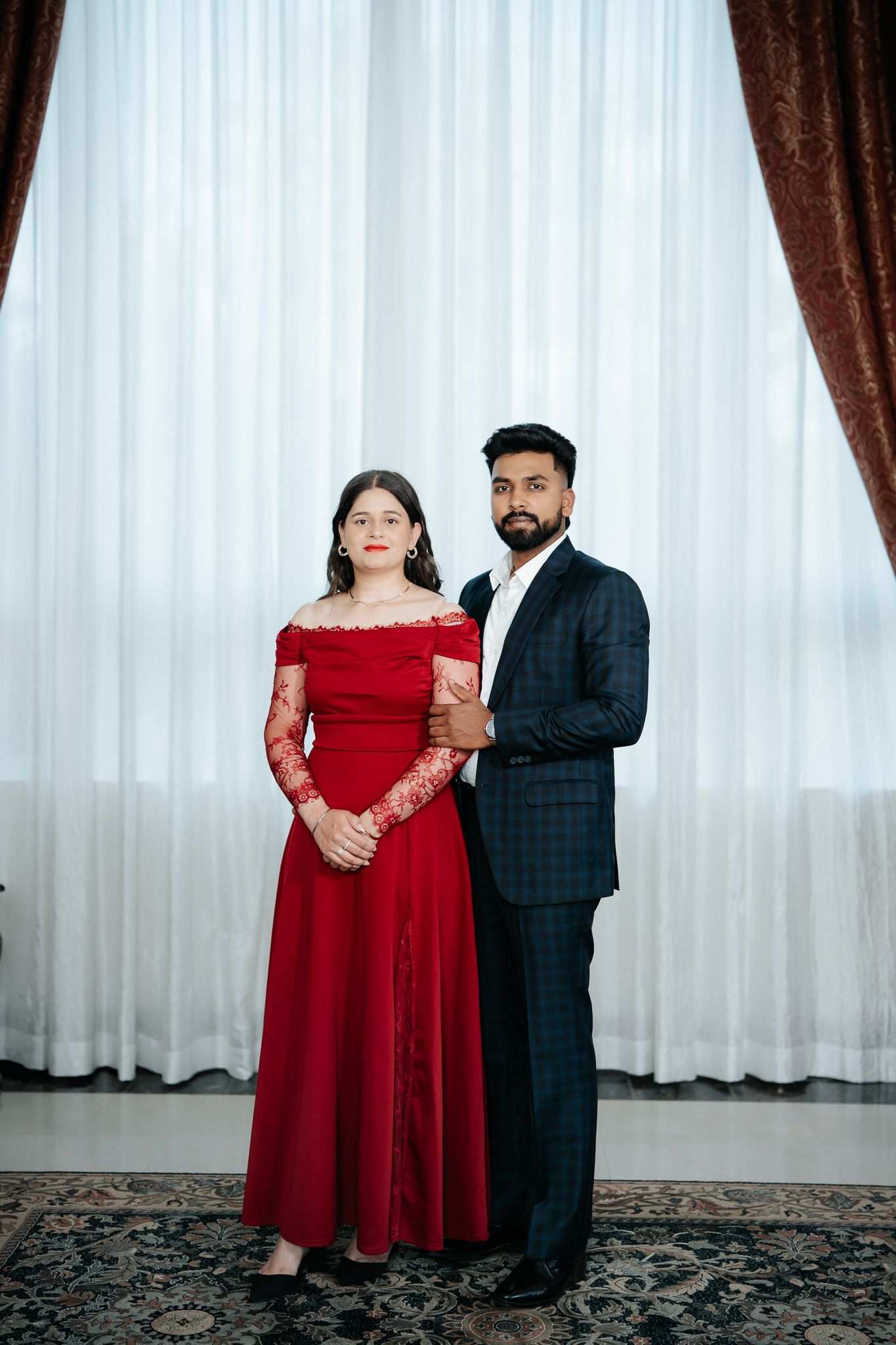 Couple posing formally, woman in red off-shoulder gown and man in plaid dark suit standing before white curtains.