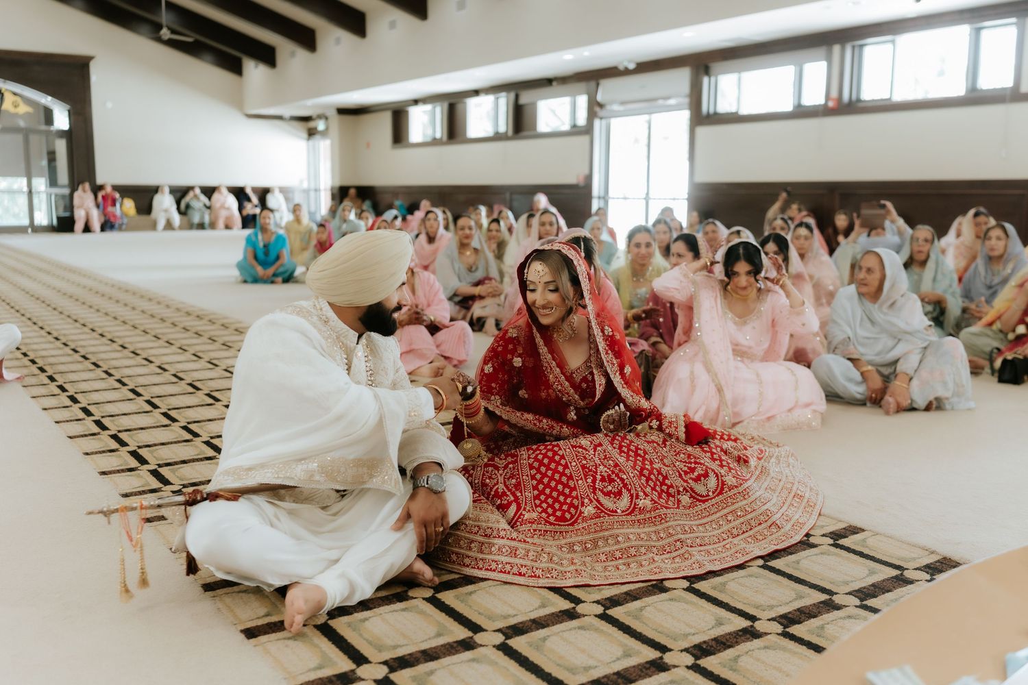 Sikh wedding ceremony with bride in red lehenga and groom in white sitting on floor in decorated hall.