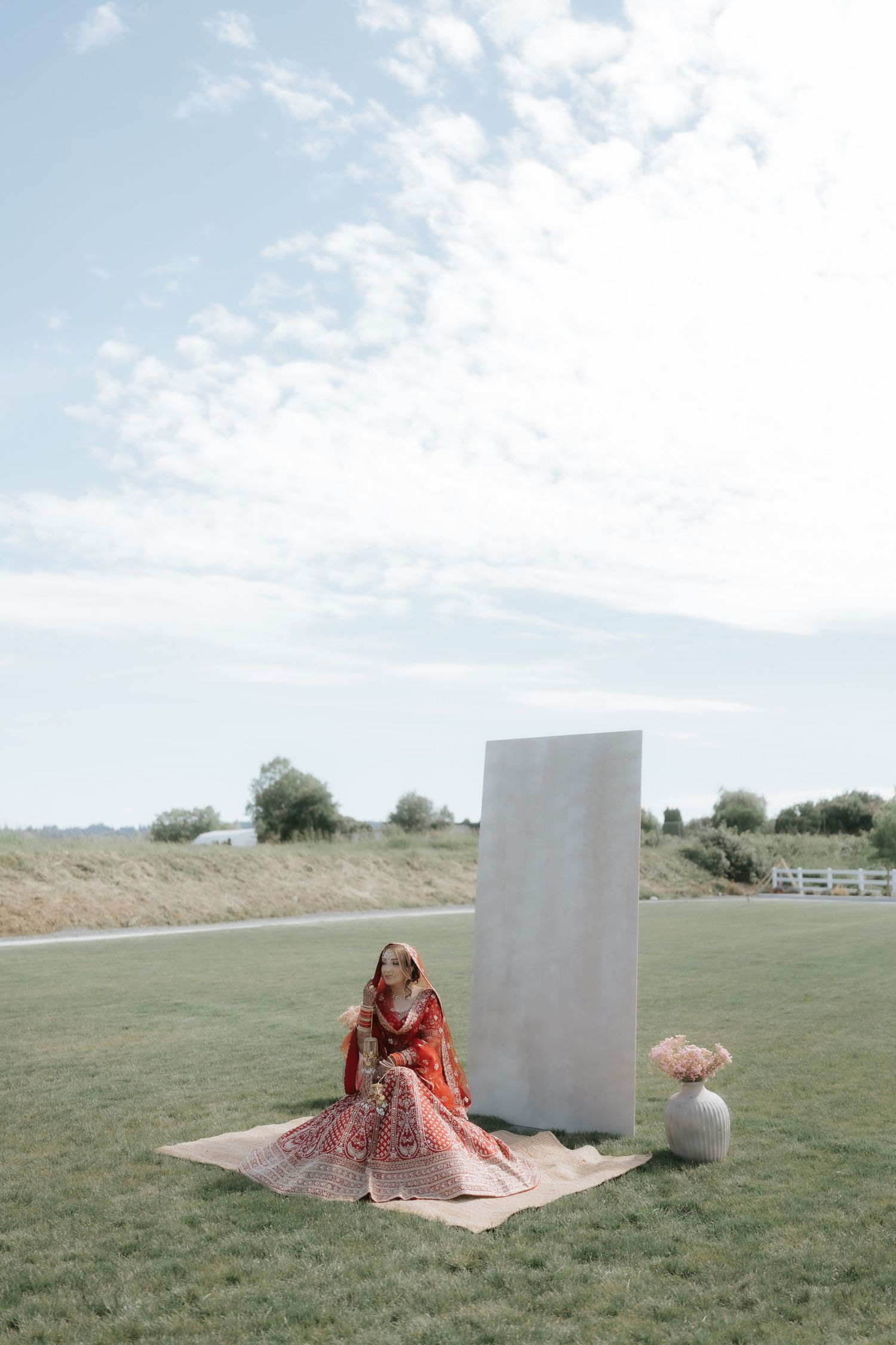 Bride in red lehenga sitting on mat outdoors beside white panel and vase under cloudy sky.