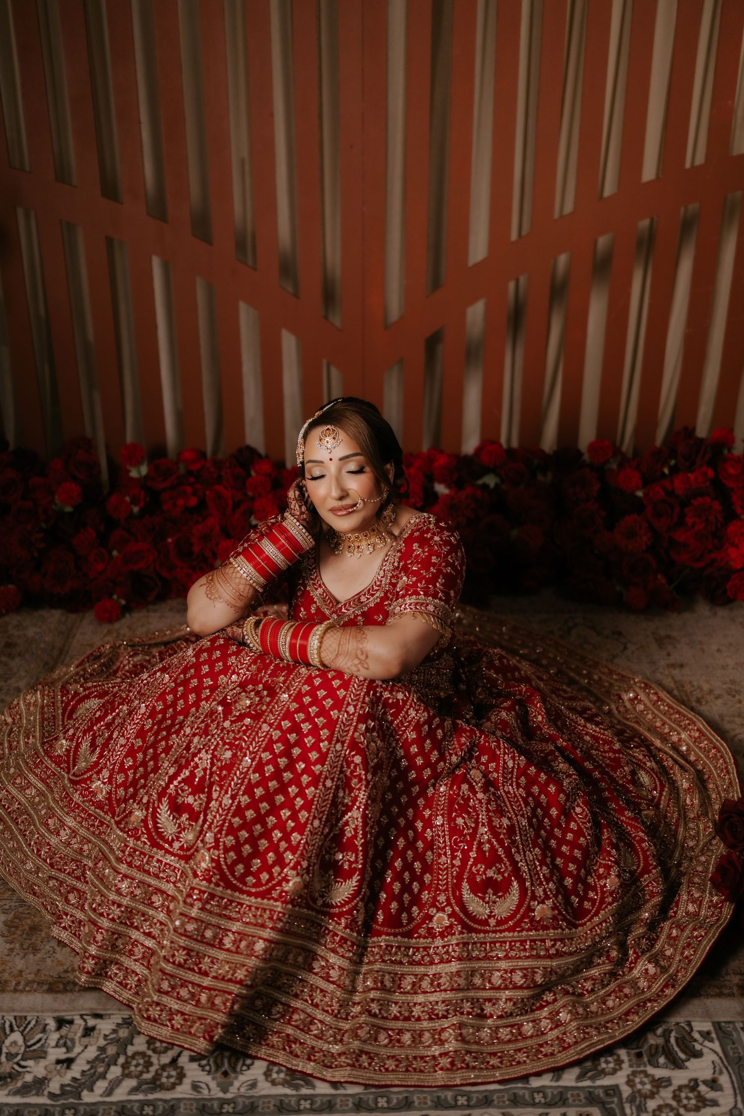 Indian bride in red embroidered lehenga with gold jewelry sits gracefully on floor, showcasing traditional bridal attire.