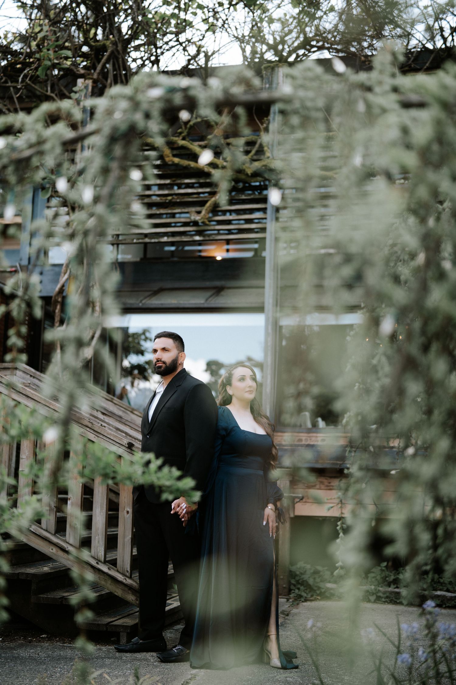 Couple in formal attire standing under a rustic pergola surrounded by lush greenery at dusk.