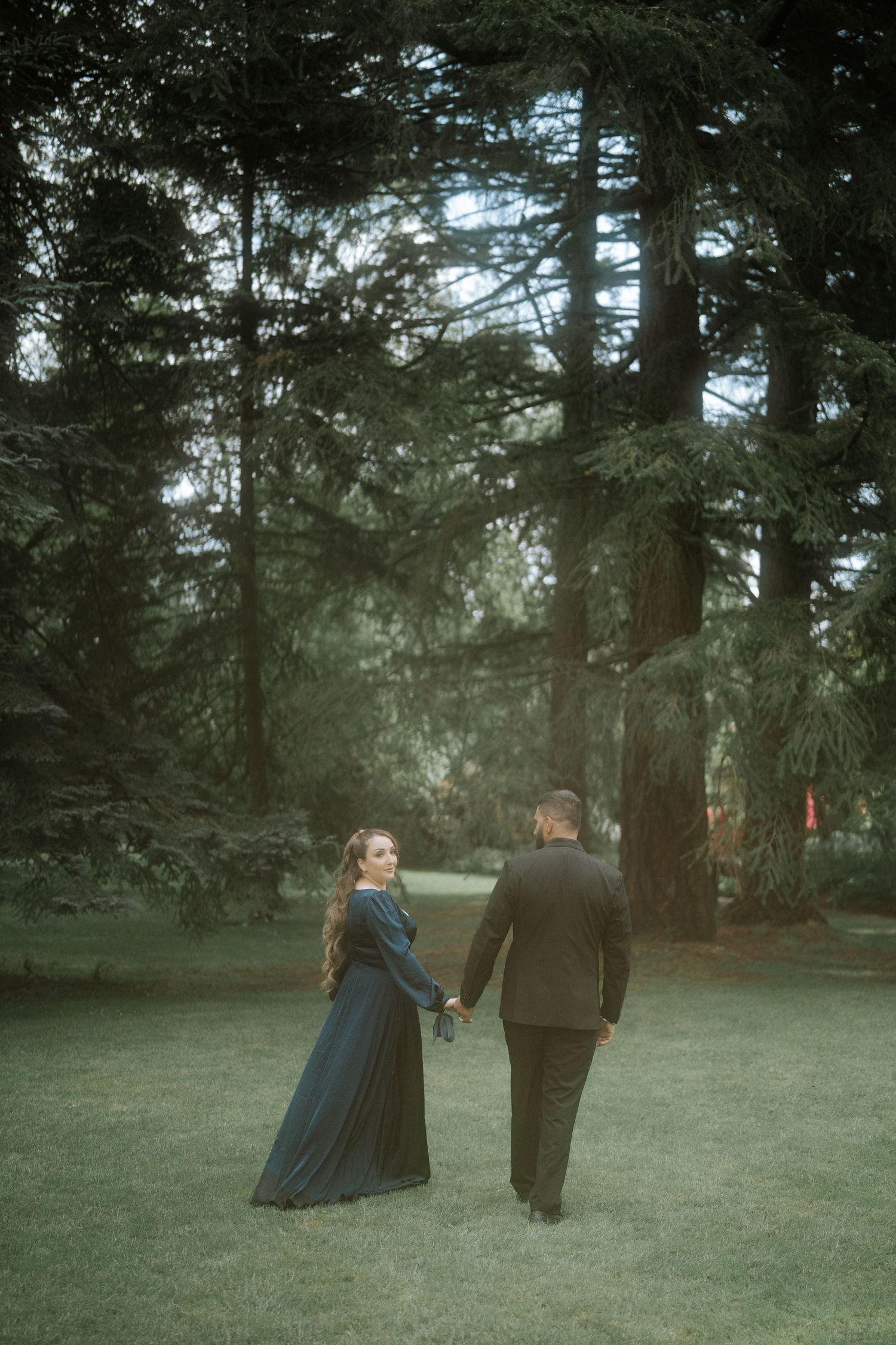 Couple in formal attire holding hands while walking through a lush green forest park at dusk.