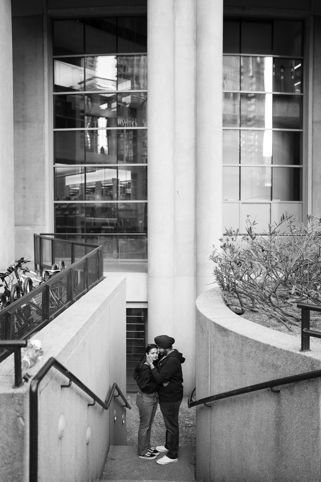 A black and white photograph of two people embracing in a stairwell of a modern building with glass windows.