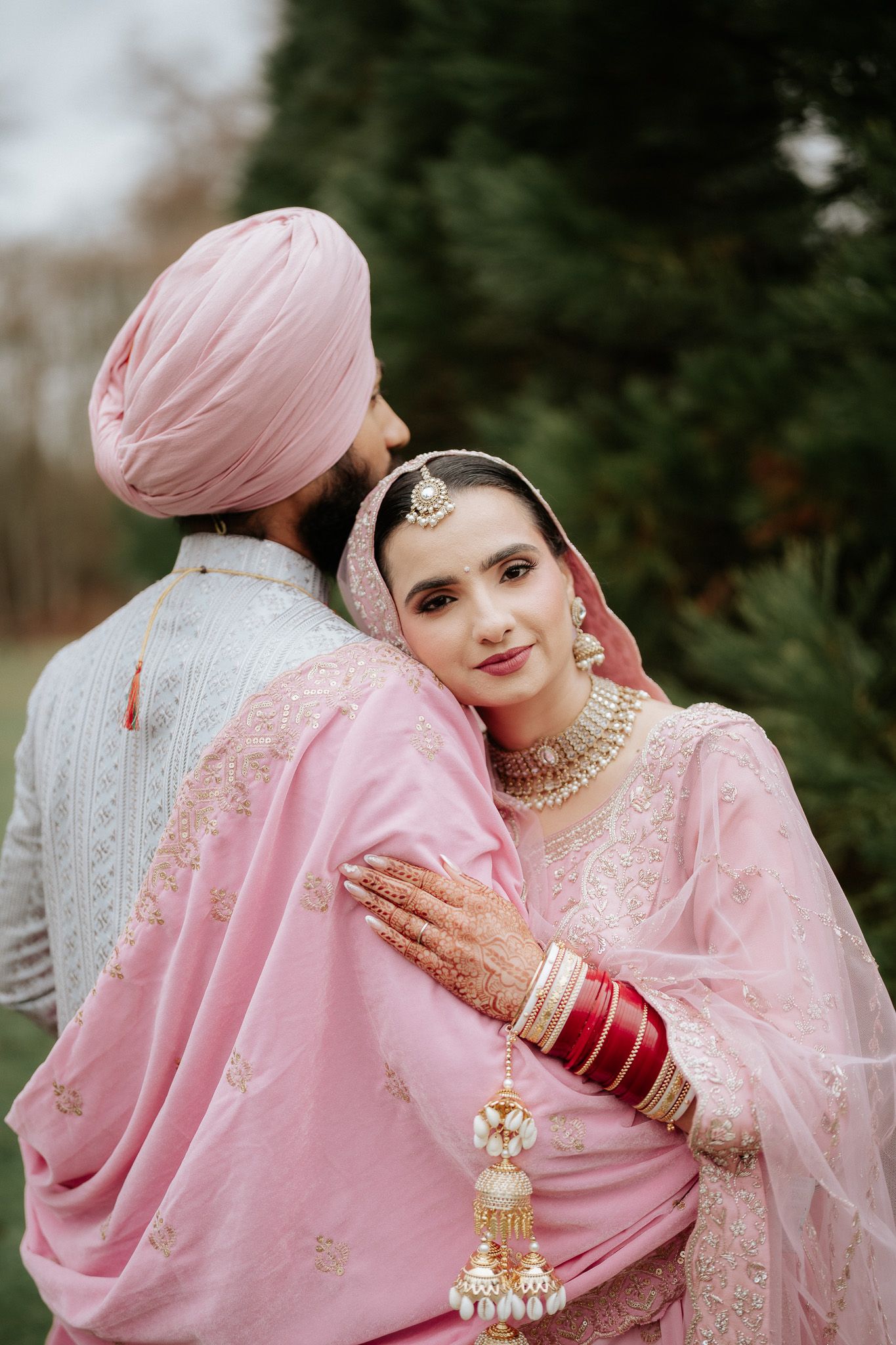 A couple in traditional Sikh wedding attire embrace while wearing pink and silver outfits with ornate jewelry and decorations.