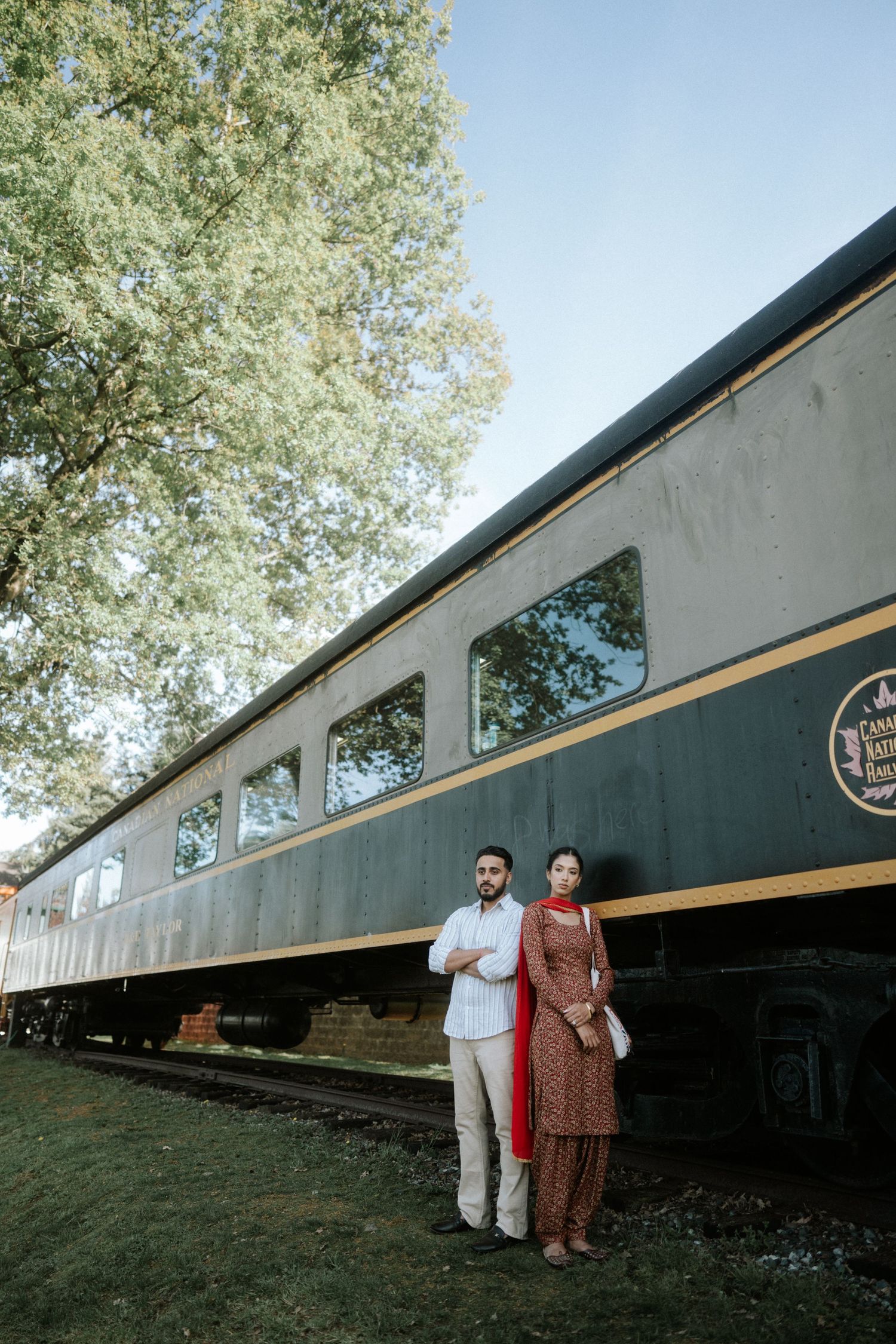 A couple poses in front of a vintage green and white train car on a sunny day with trees in the background.