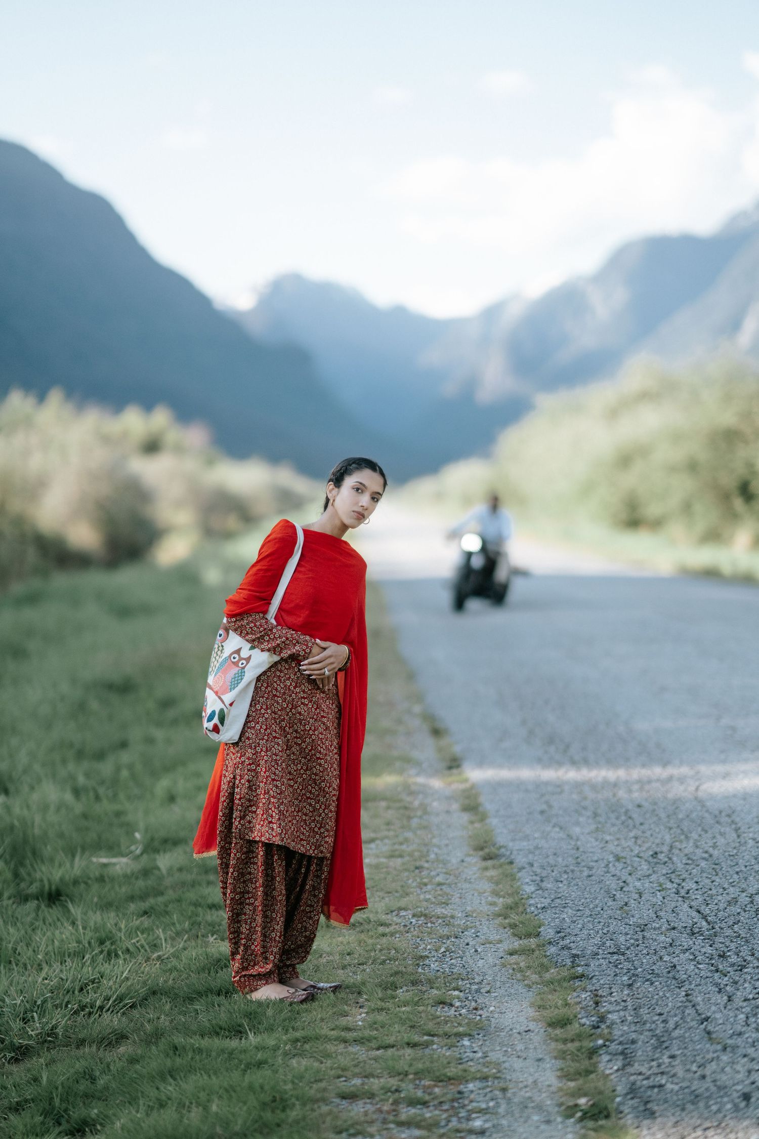 Person in red sweater and patterned skirt walks along mountain road on misty day.