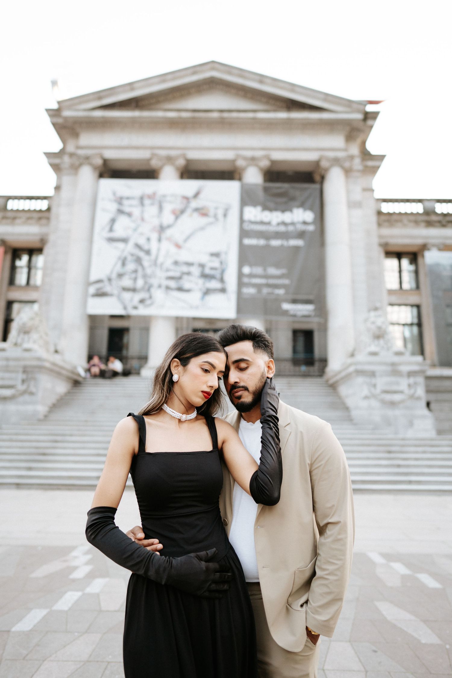 A couple in elegant attire share an intimate moment in front of a grand neoclassical building with white columns.