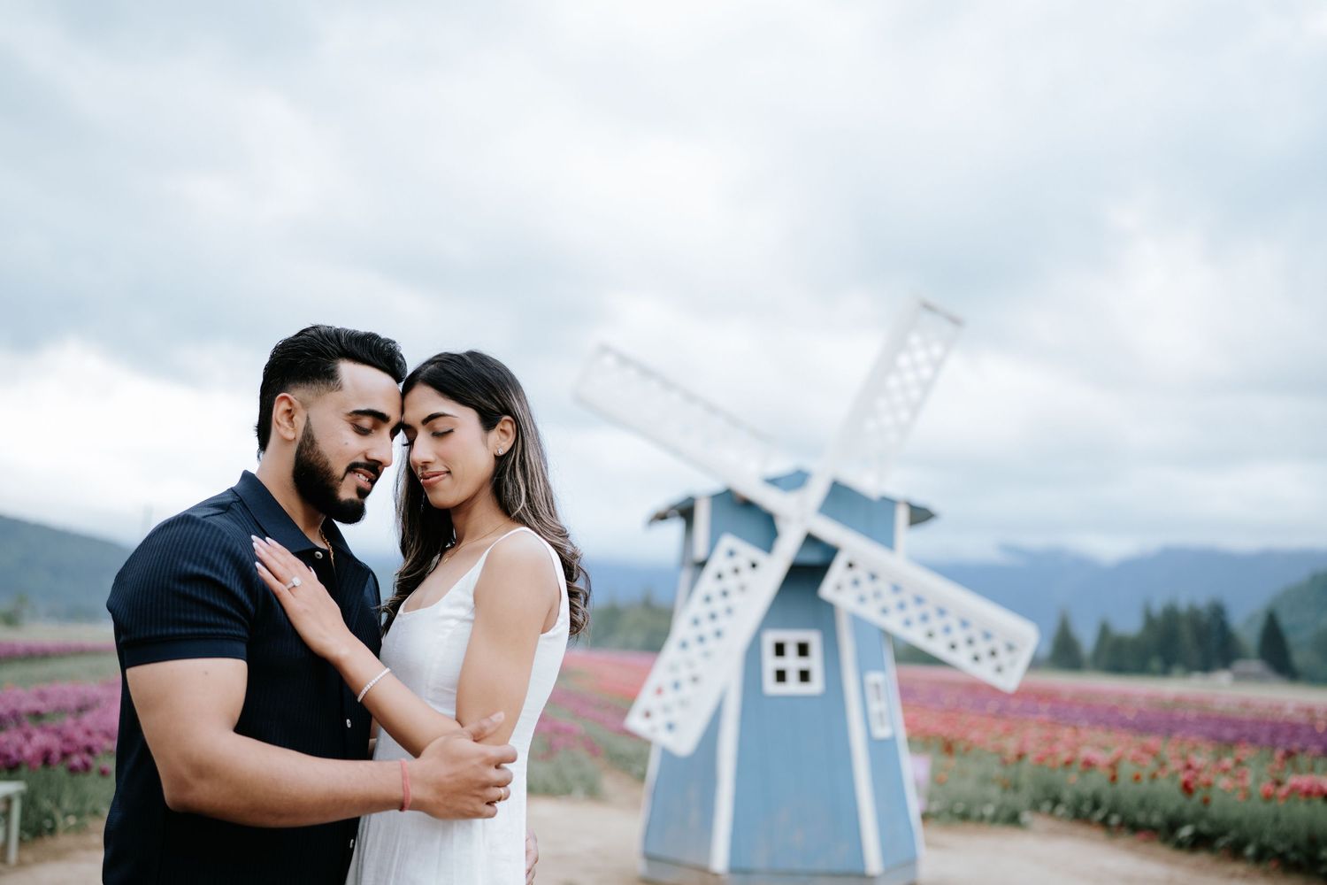 A couple embraces in front of a blue windmill with mountains and a tulip field in the background.