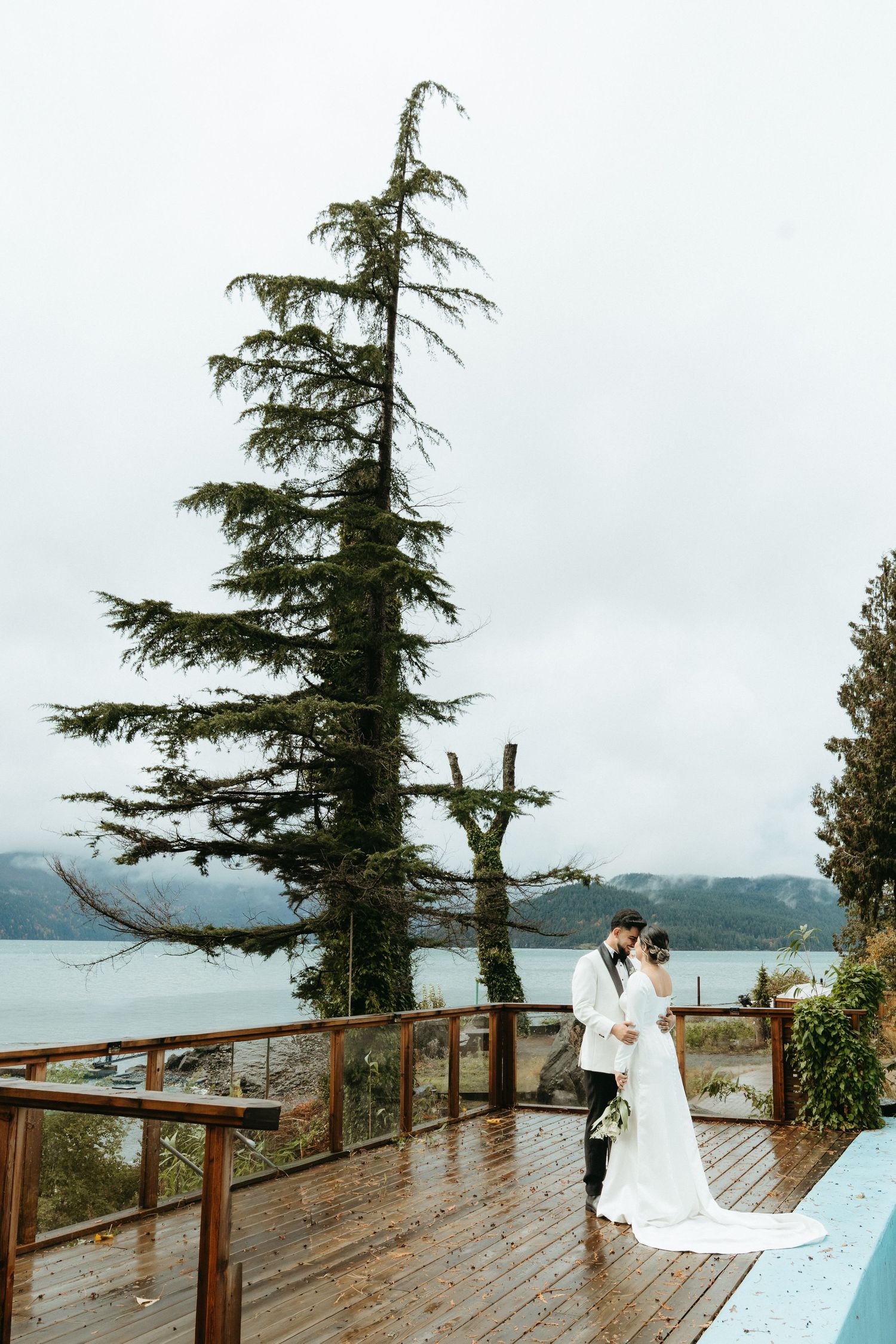A couple shares an intimate moment on a wooden deck overlooking a misty lake surrounded by tall evergreen trees.