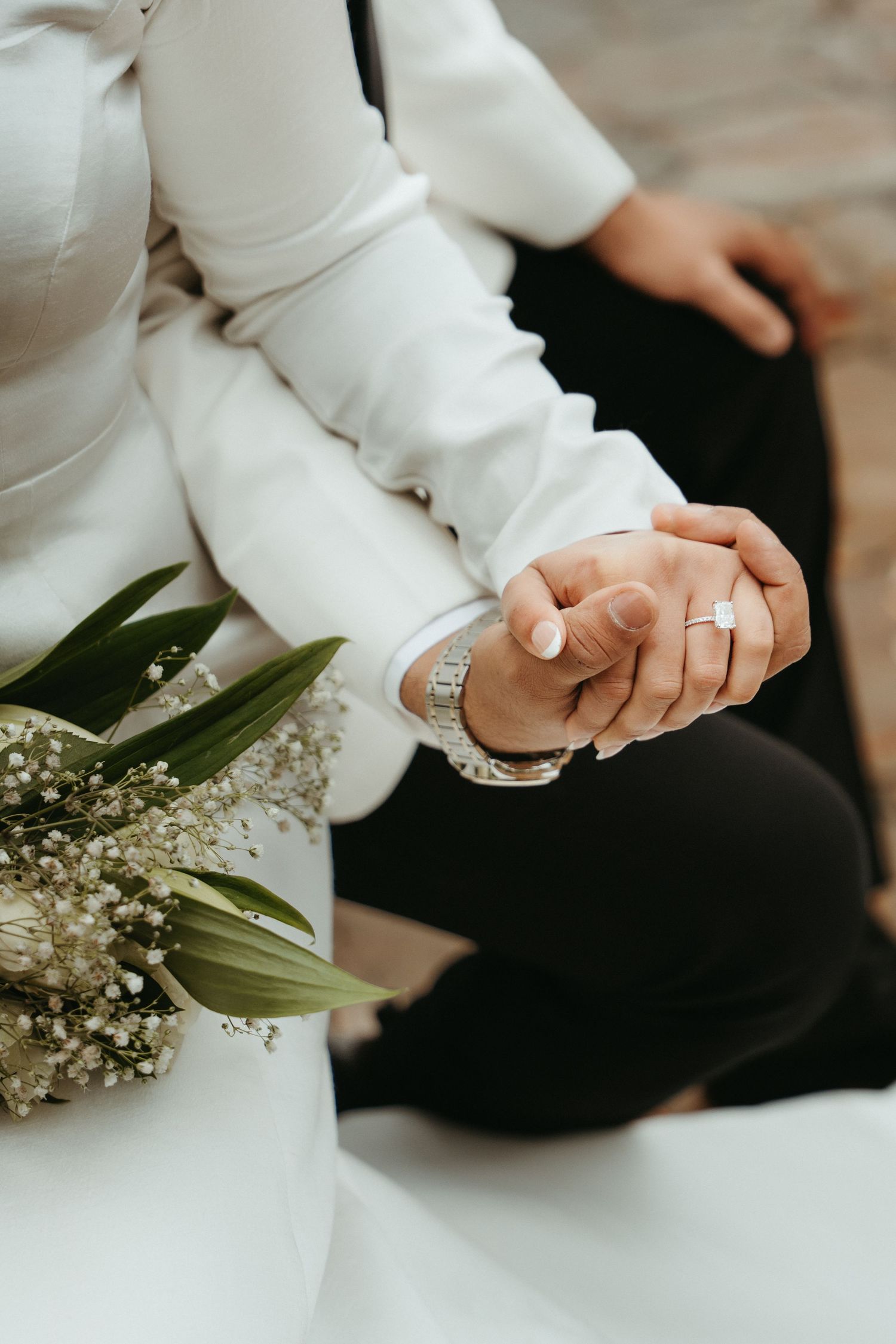 Close-up of clasped hands with wedding rings and white baby's breath flowers against formal white and black attire.