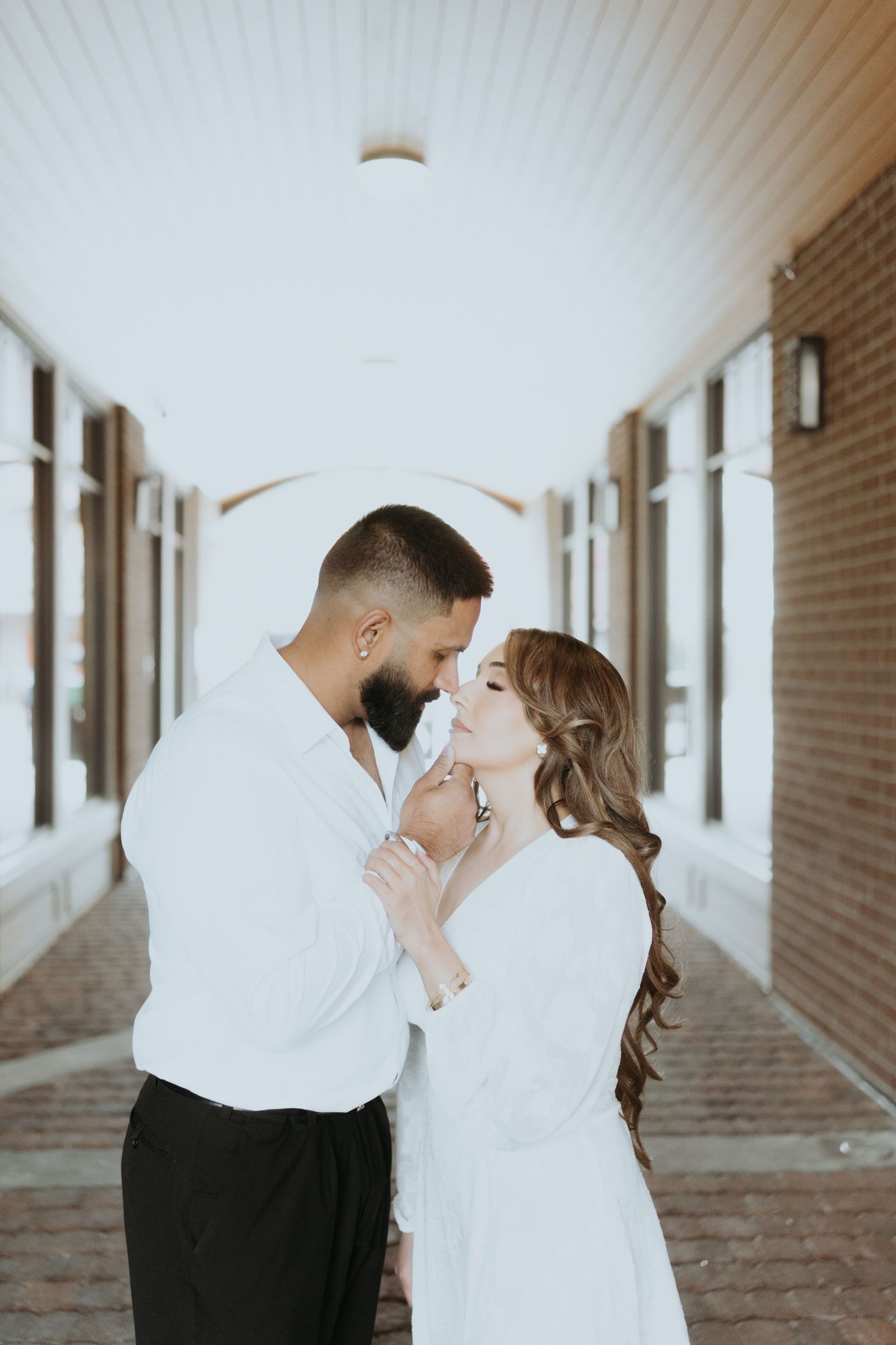 Romantic couple poses in a sunlit brick corridor with white ceiling panels.