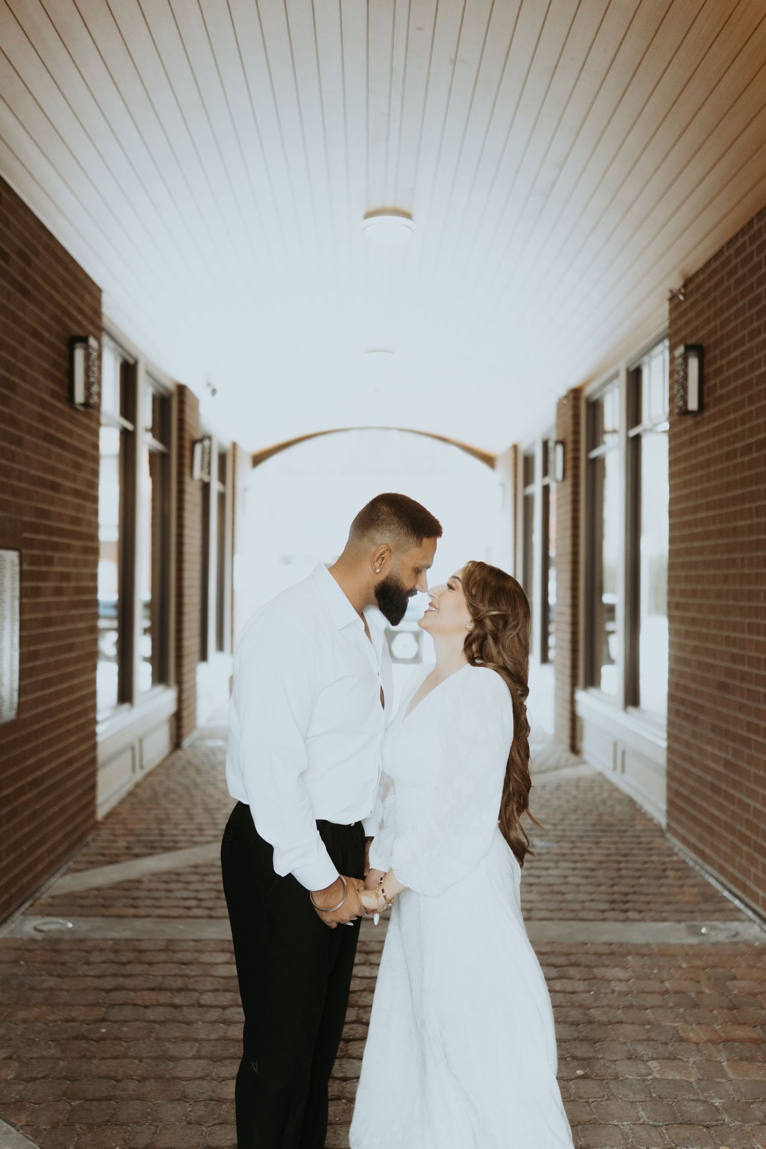 A couple in white clothing embraces in a brick corridor with natural lighting.