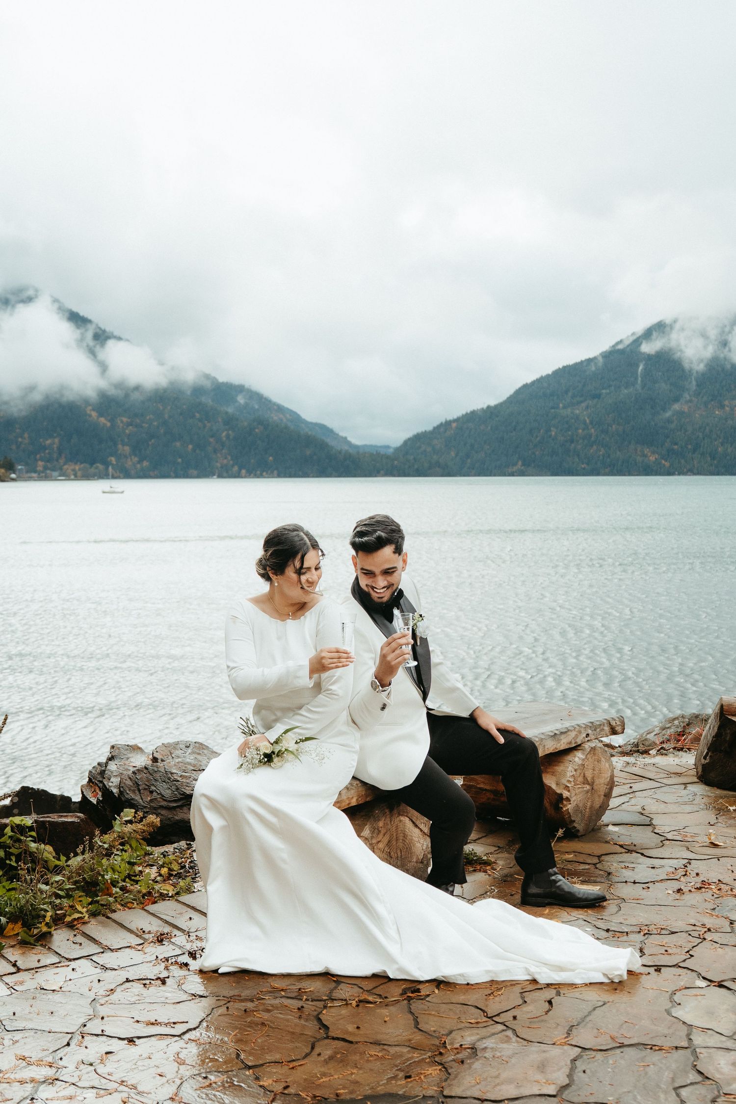 A couple in wedding attire sits together on rocky shoreline with misty mountain lake views in the background.