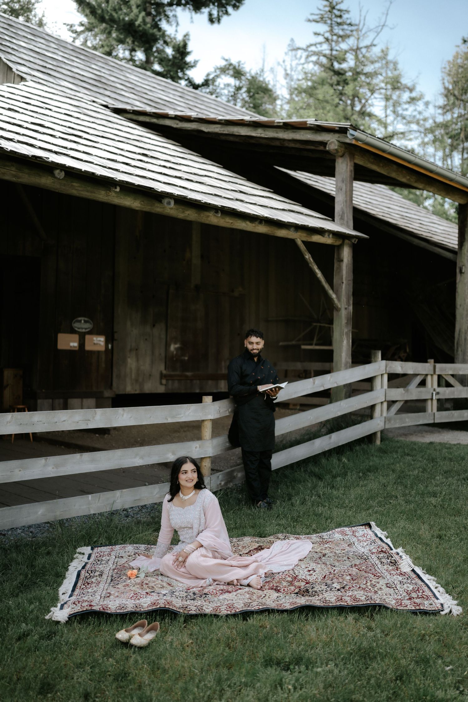 A couple relaxes by a rustic barn, with one person seated on a decorative rug outdoors.