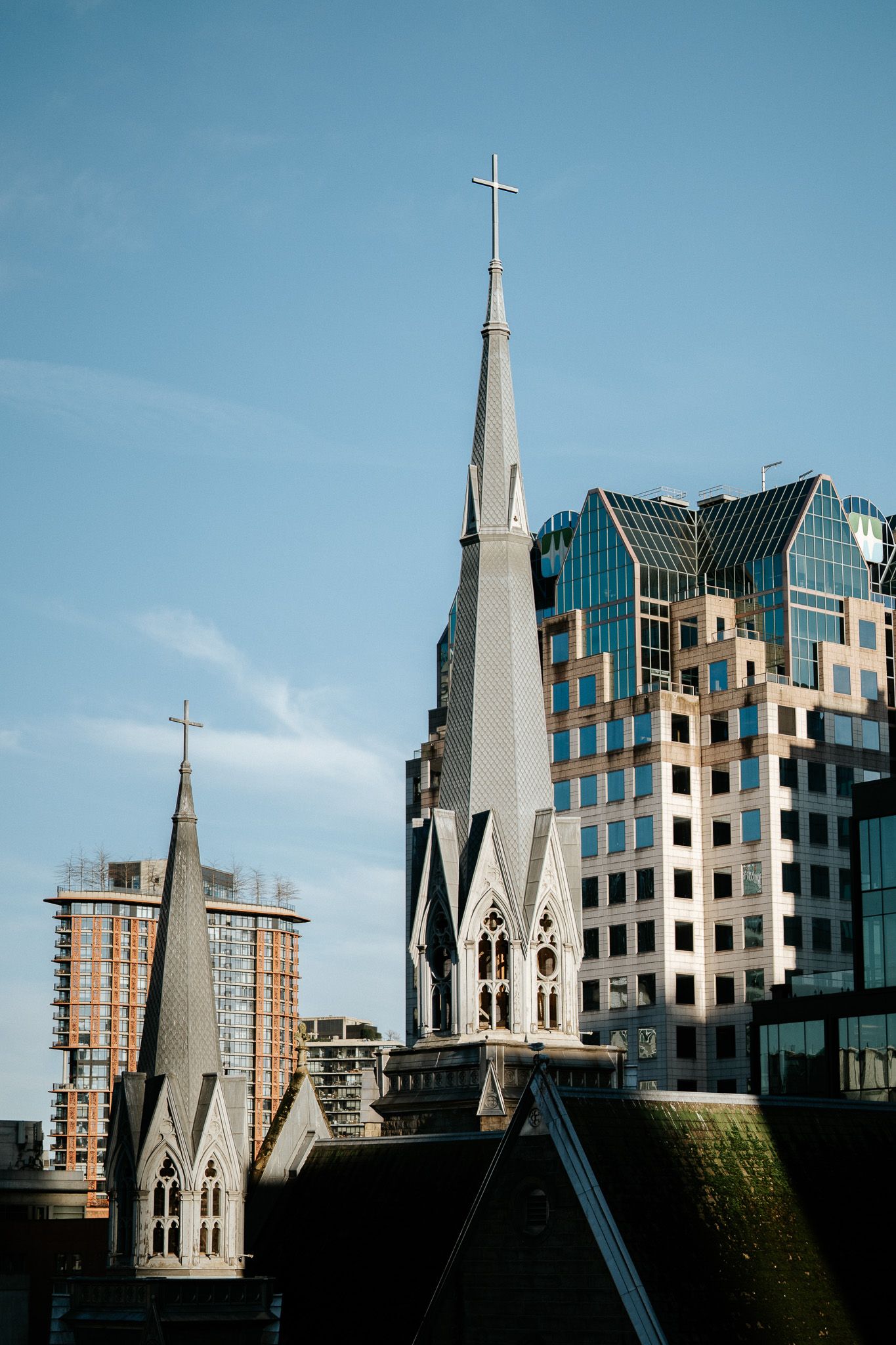 Modern church spire with angular architecture rises against blue sky next to contemporary glass office buildings.