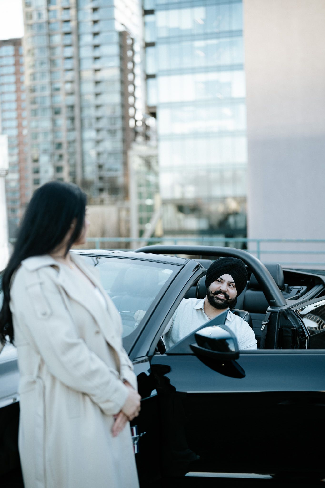 A person in a white coat walks toward and enters a black car on a city street.