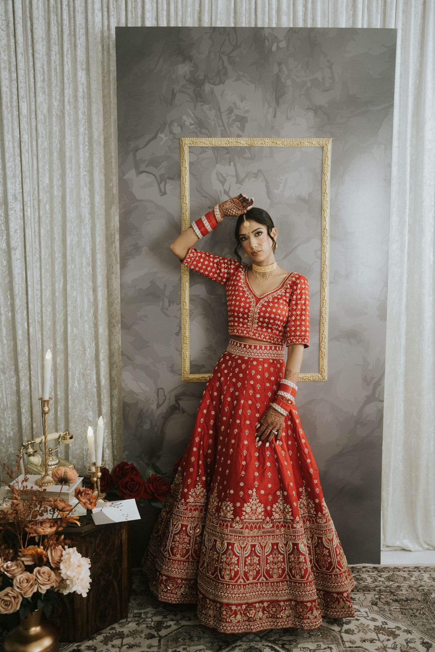 Elegant red bridal lehenga showcased in a studio setting with gold frames and floral arrangements.