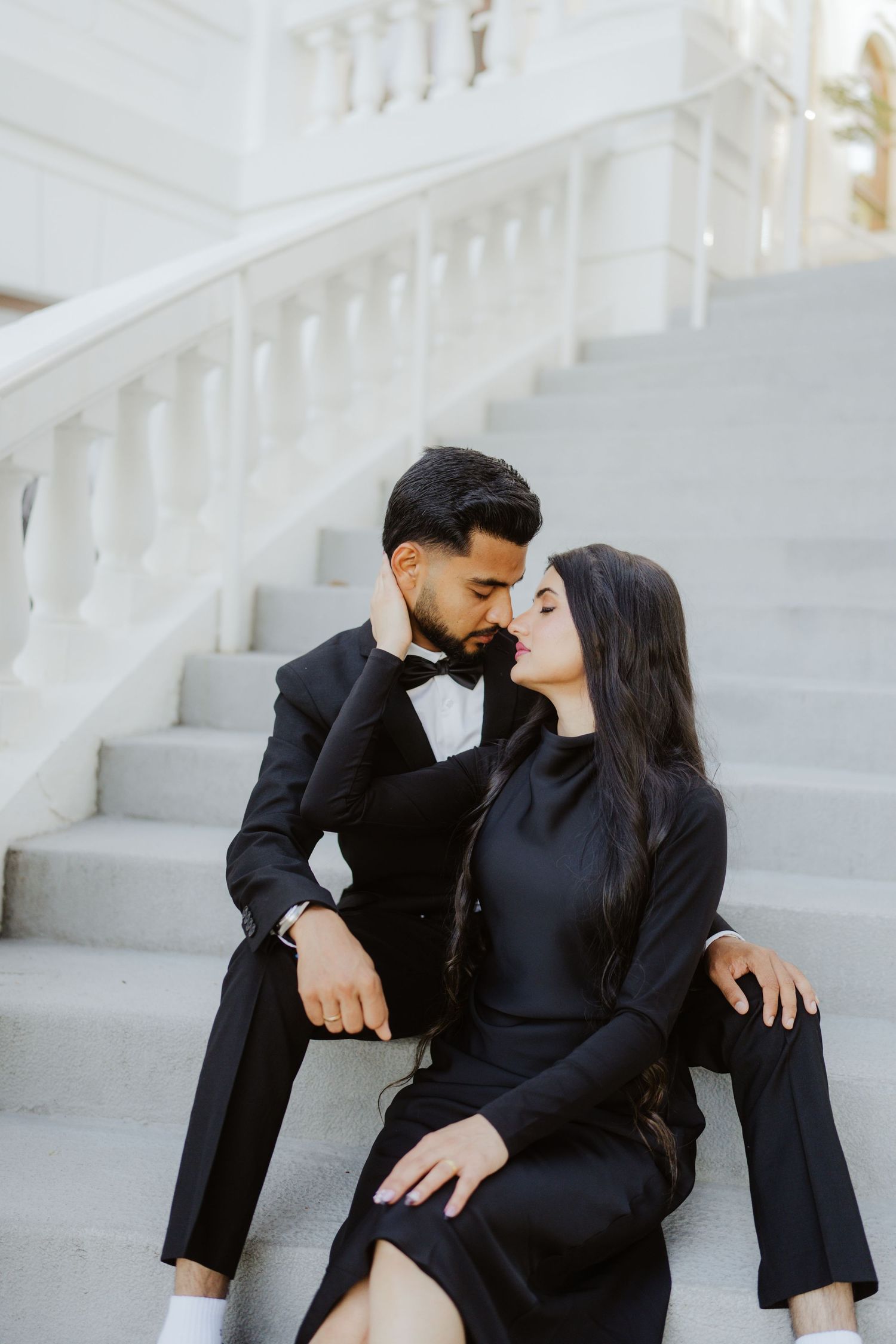 A couple in elegant black formal attire share a romantic moment on white marble steps.