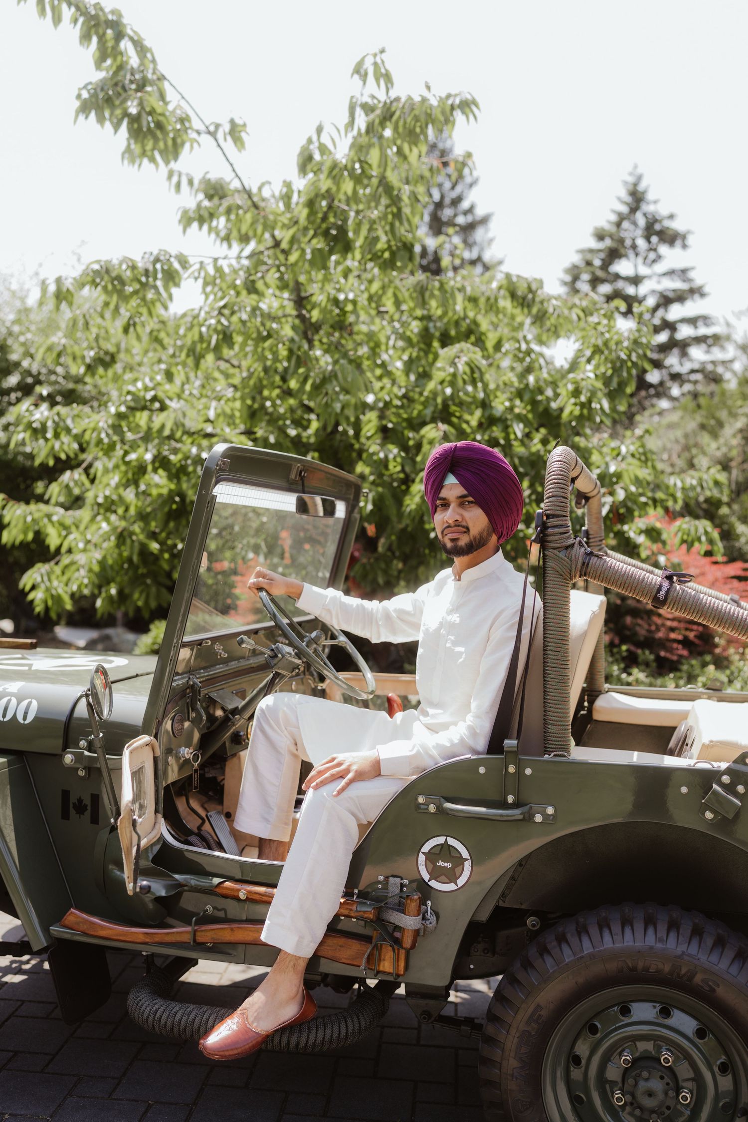 A Sikh man in a white kurta and purple turban sits at the wheel of a vintage military jeep outdoors.