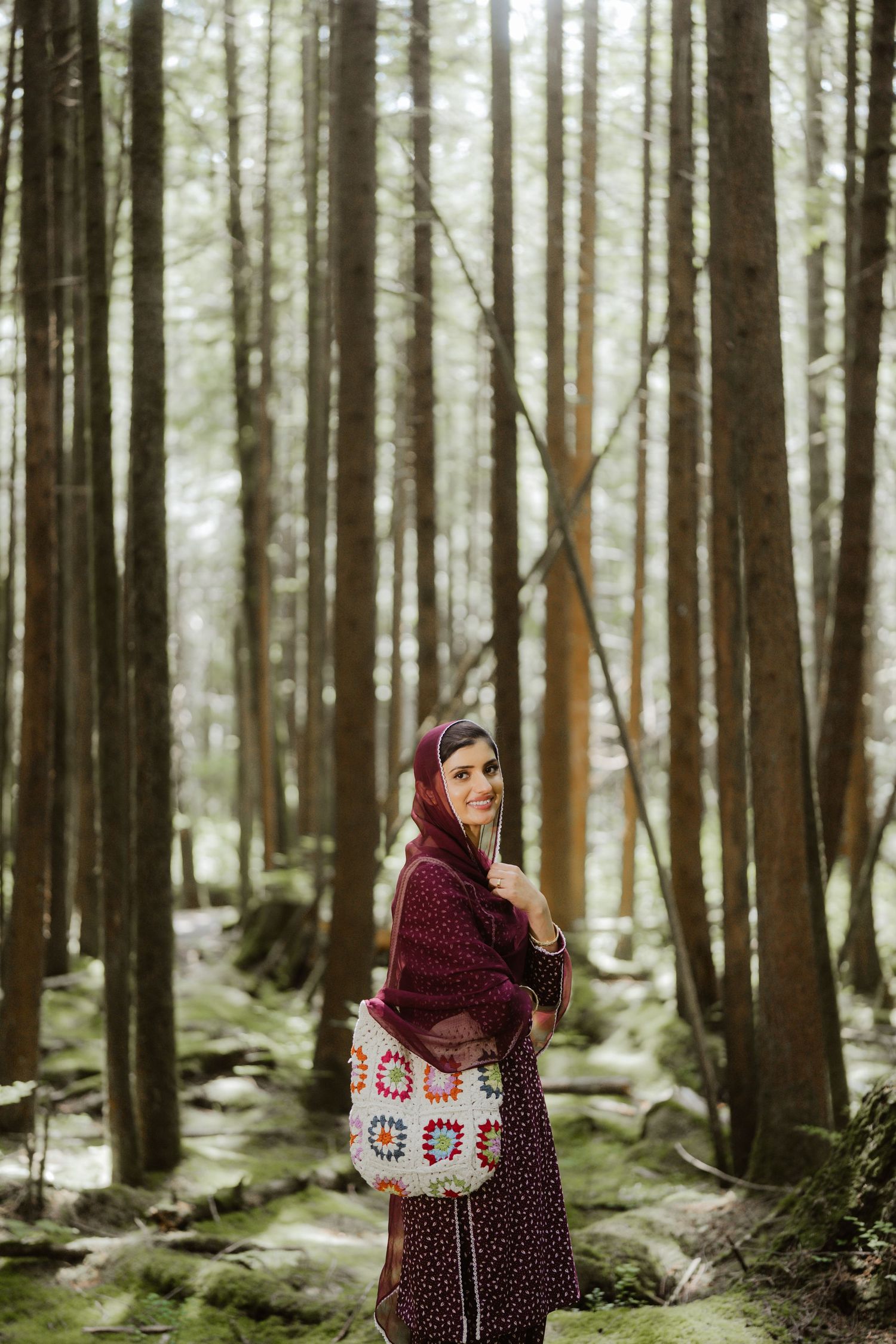 Woman in burgundy hijab and dress holding a colorful crochet bag, standing among tall trees in a misty forest.
