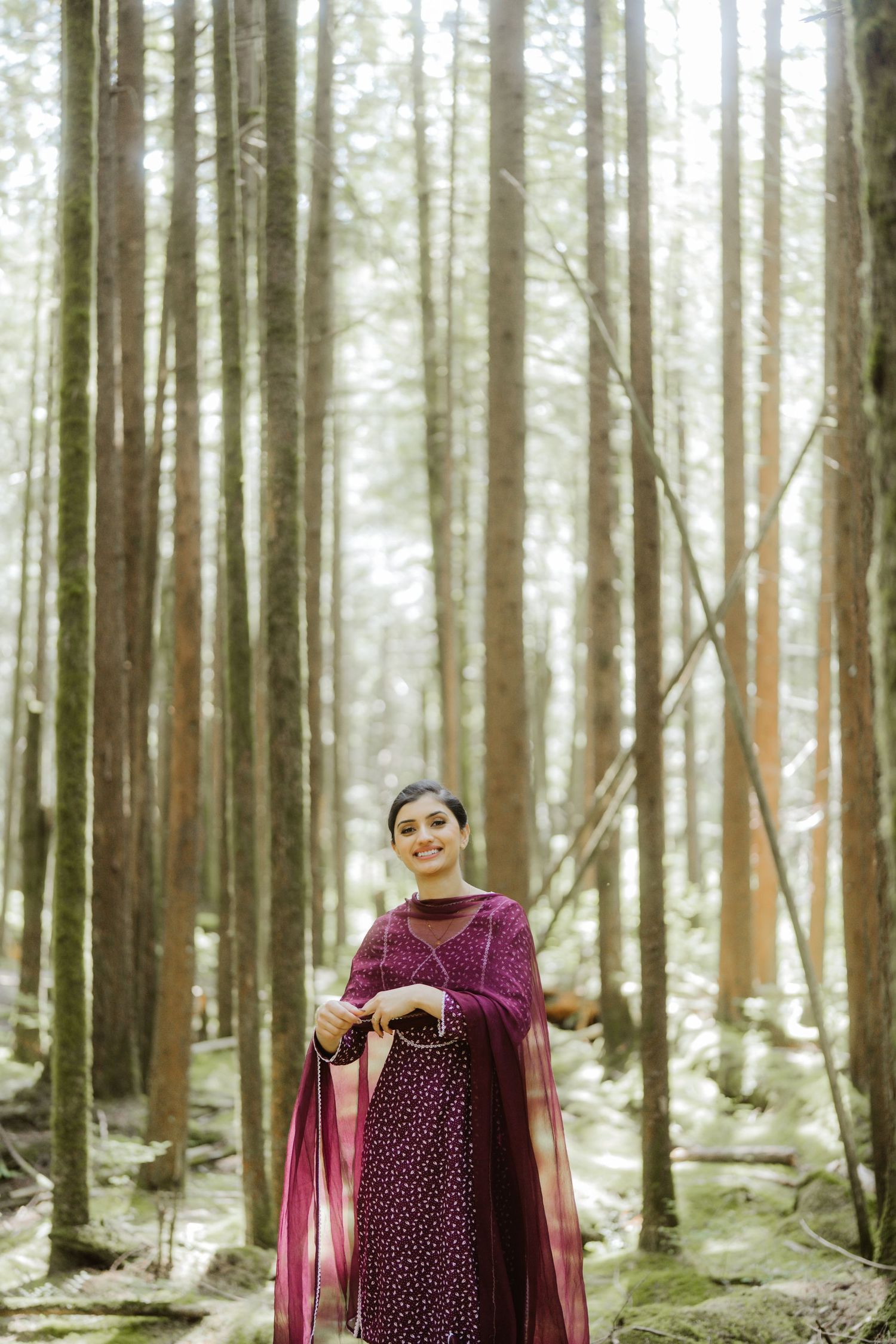 Indian woman in purple salwar kameez smiling while standing among tall trees in a serene forest.