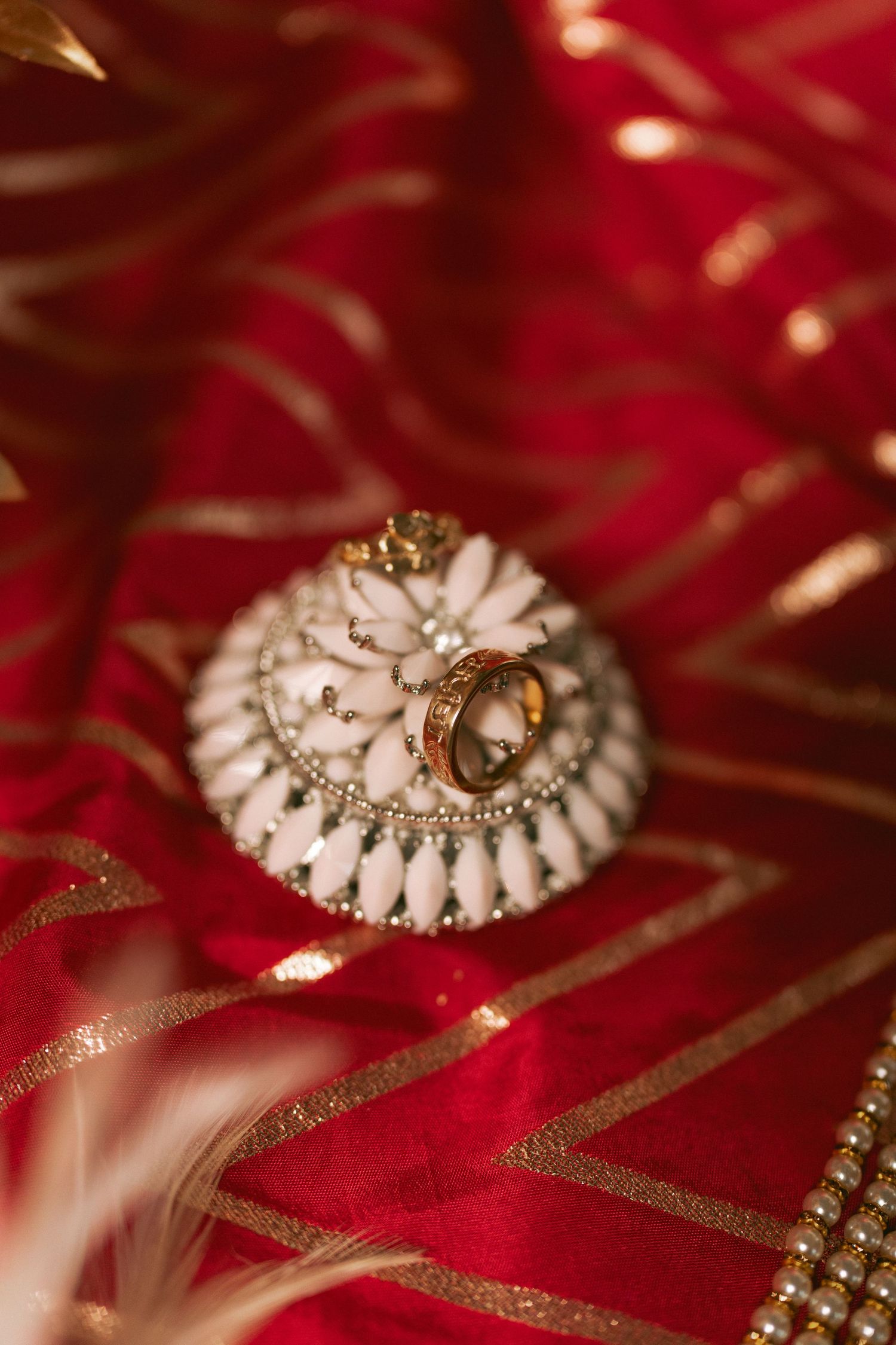 Gold wedding ring resting on white beaded jewelry piece on red and gold embroidered fabric.