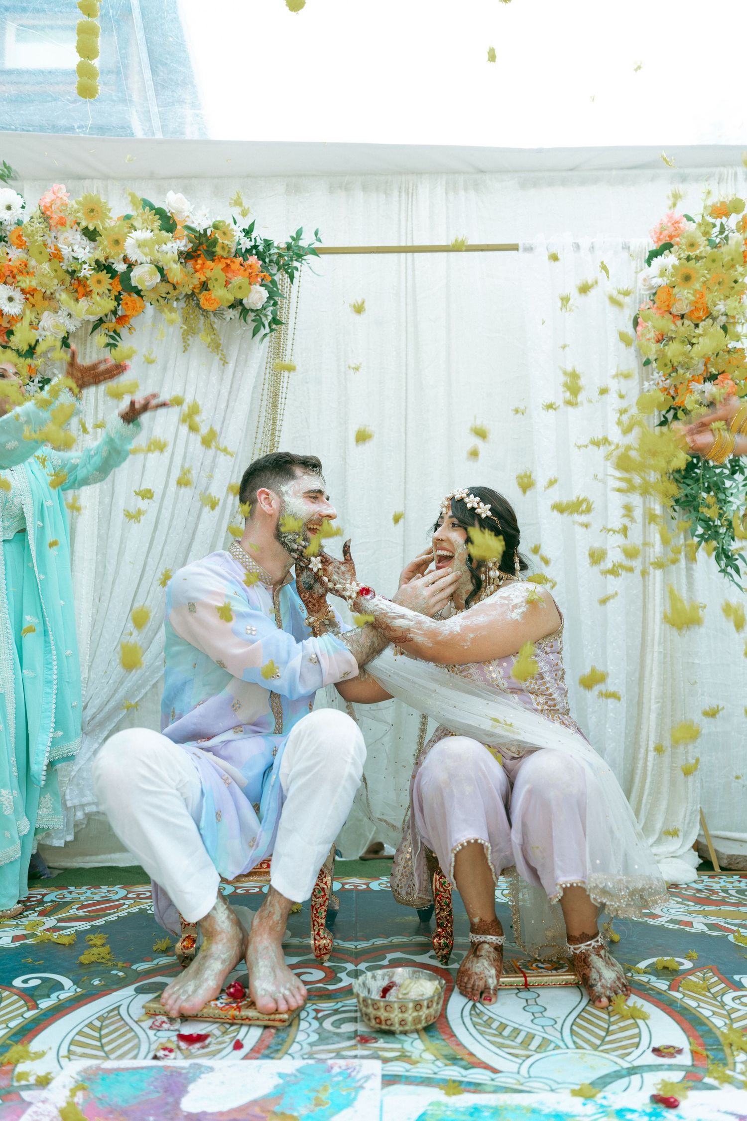 Couple laughing during Indian Haldi ceremony, surrounded by falling flower petals under floral arch with white draping.