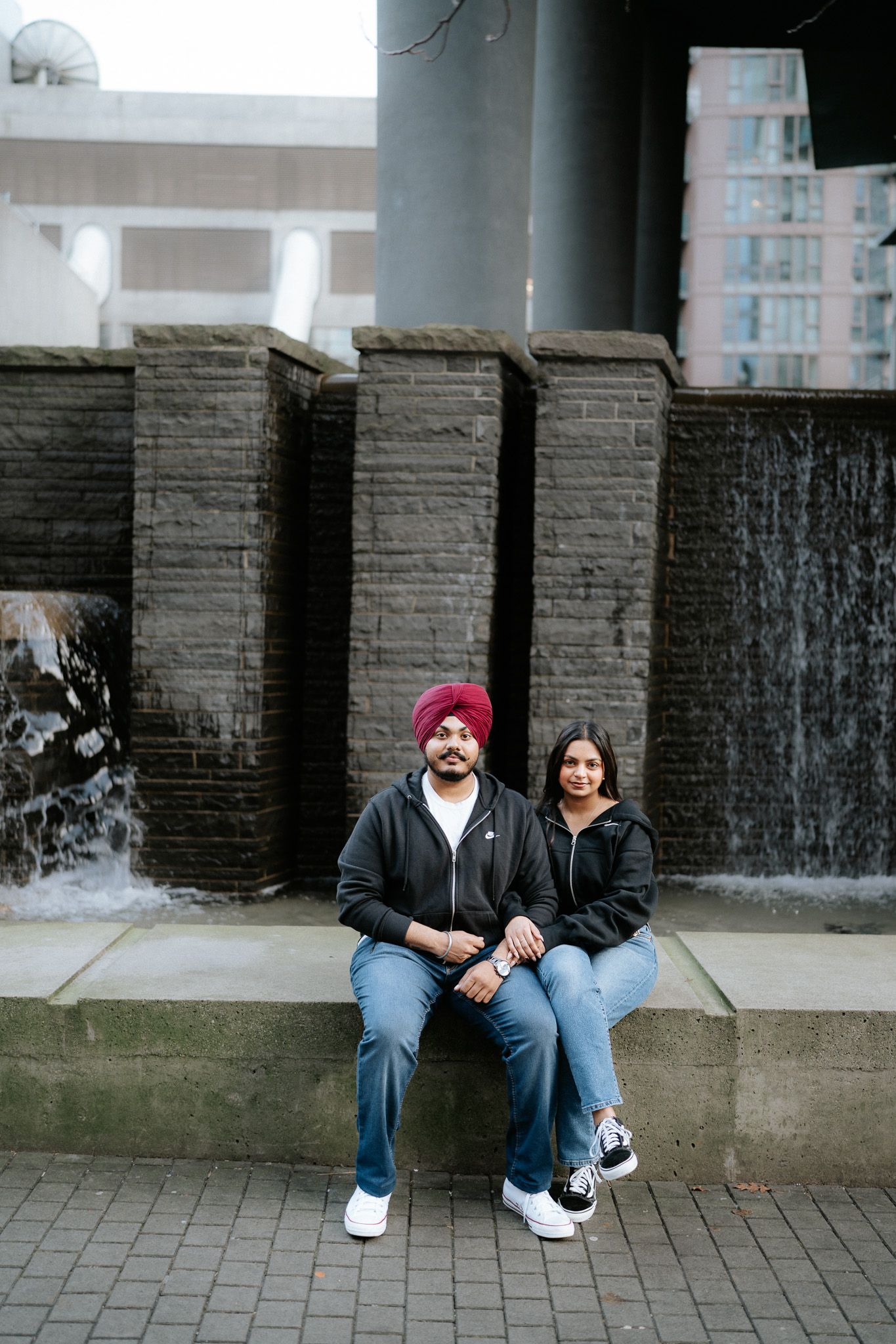 Two people sit together on concrete steps in front of a cascading water feature wall in an urban setting.