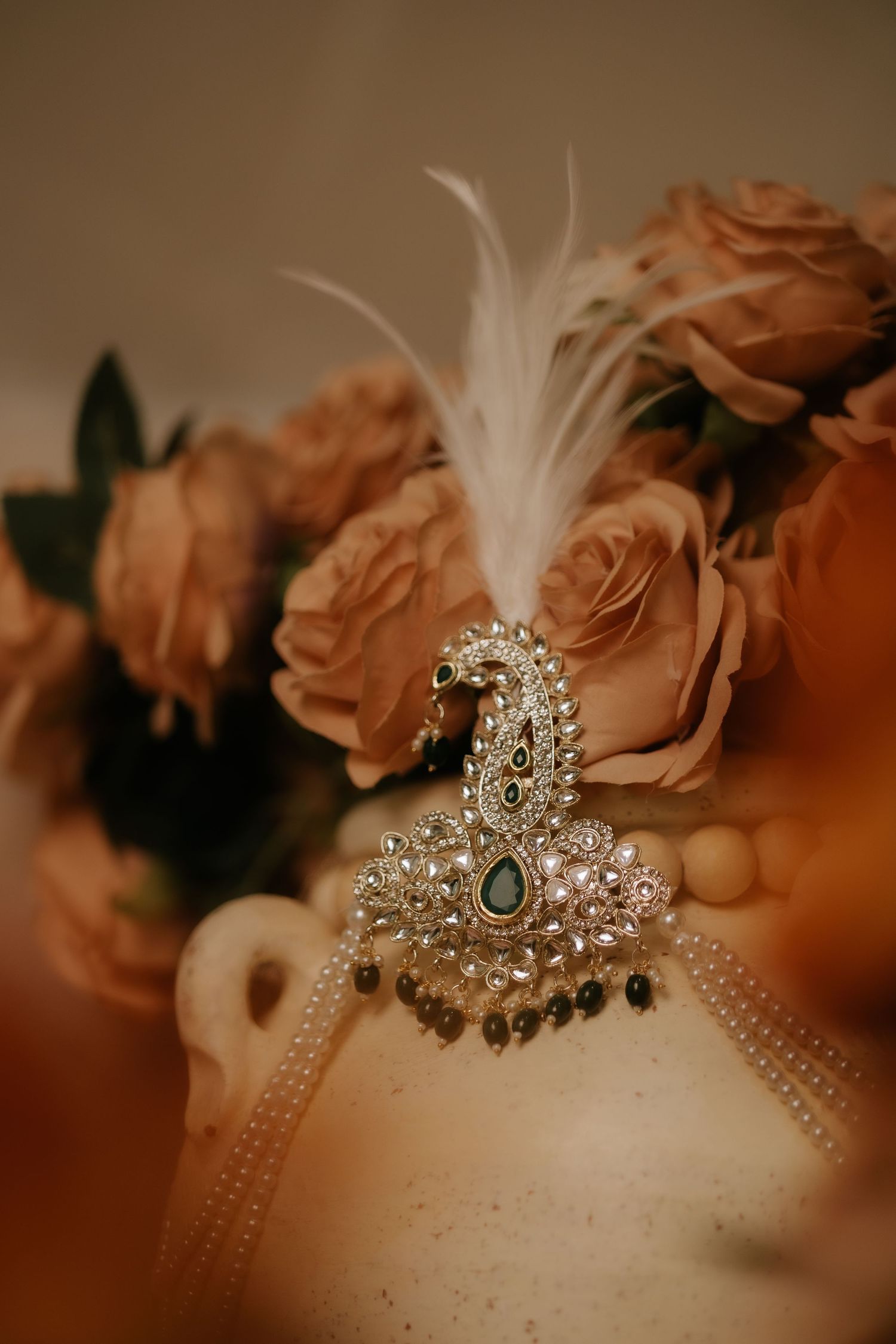 Ornate Indian peacock hair accessory with emerald gemstone, pearls, and white feather, displayed near peach roses.
